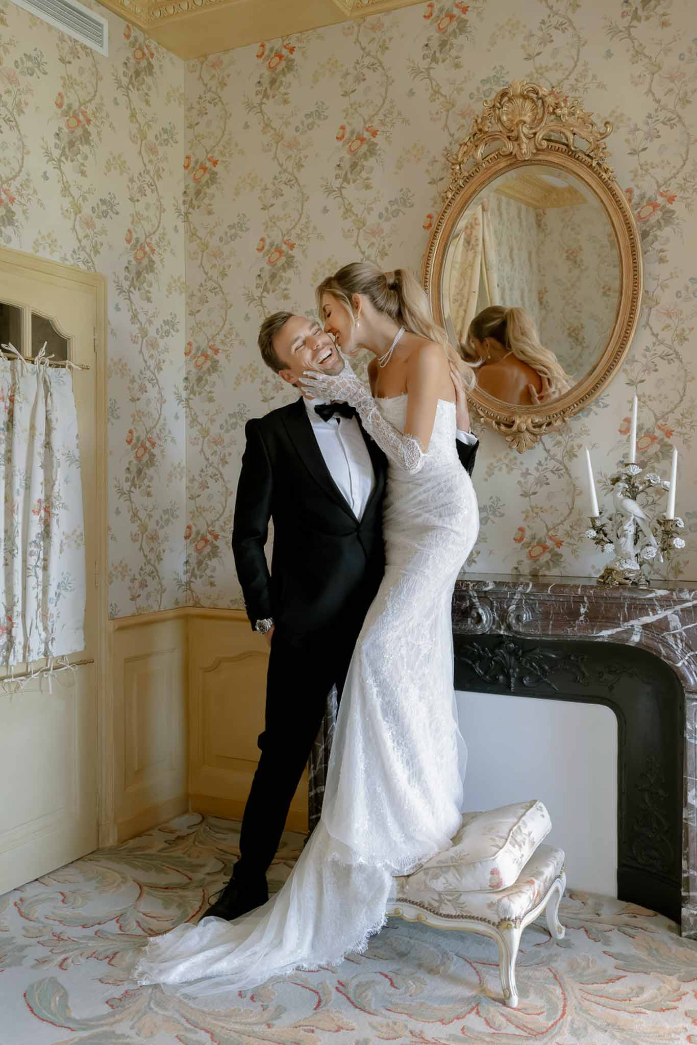 Bride kissing grooms cheek in lace gown with opera gloves in chateau room with floral wallpaper marble fireplace and gilt ...