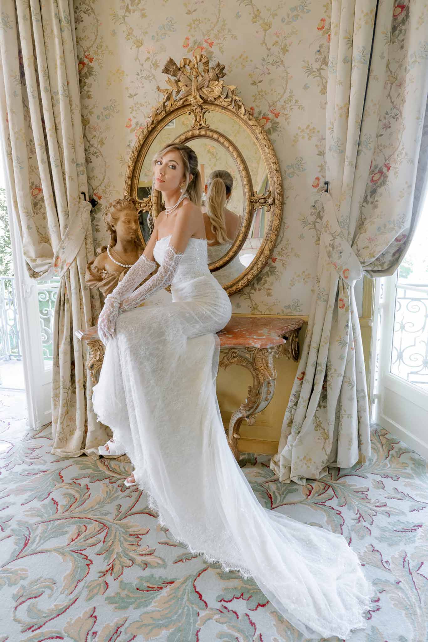 Bride in strapless lace gown with lace gloves and pearl necklaces perched on gilded console before oval mirror in chateau ...