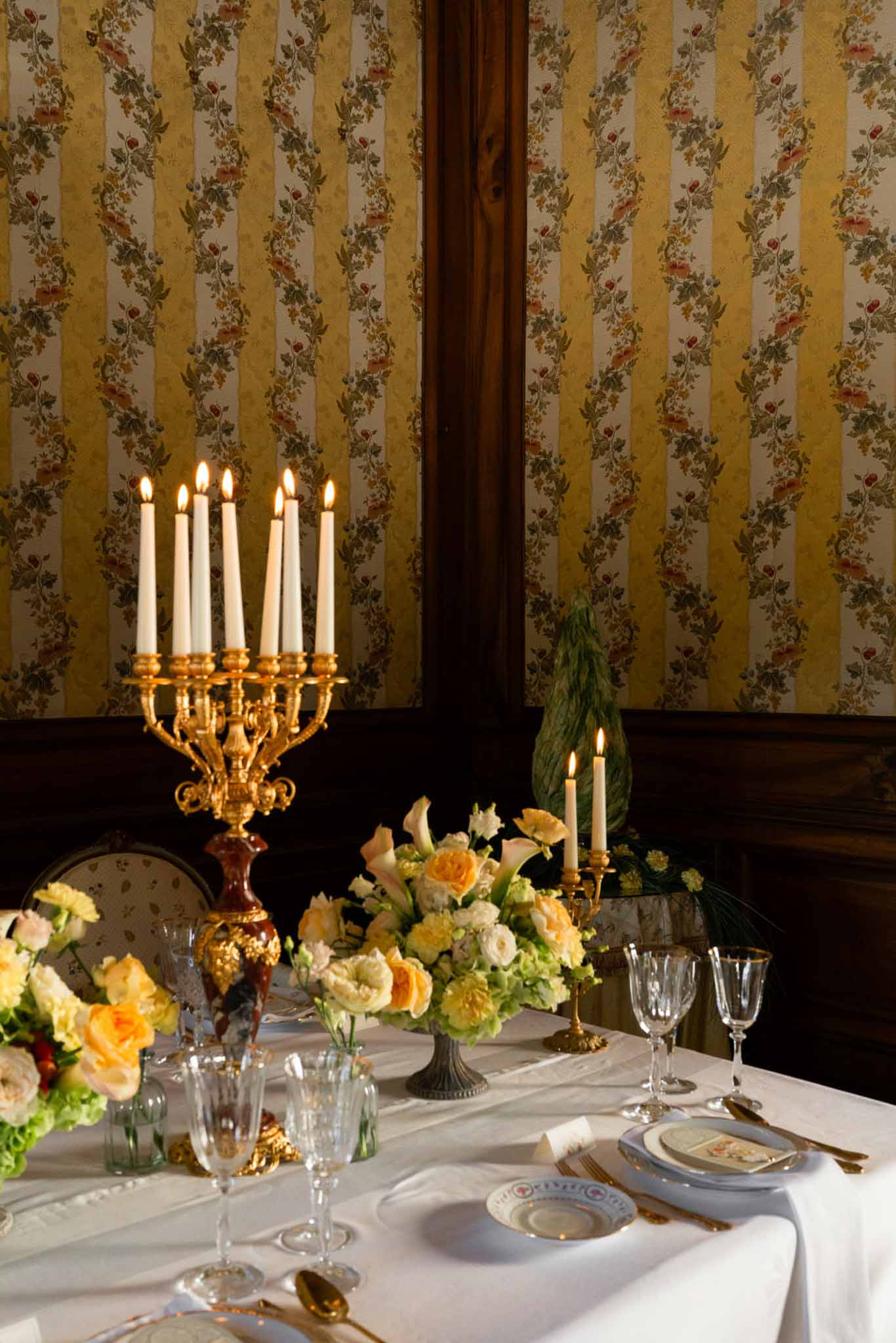 Reception table with gilt candelabra, peach and yellow roses, crystal glassware, and gold cutlery in a chateau dining room