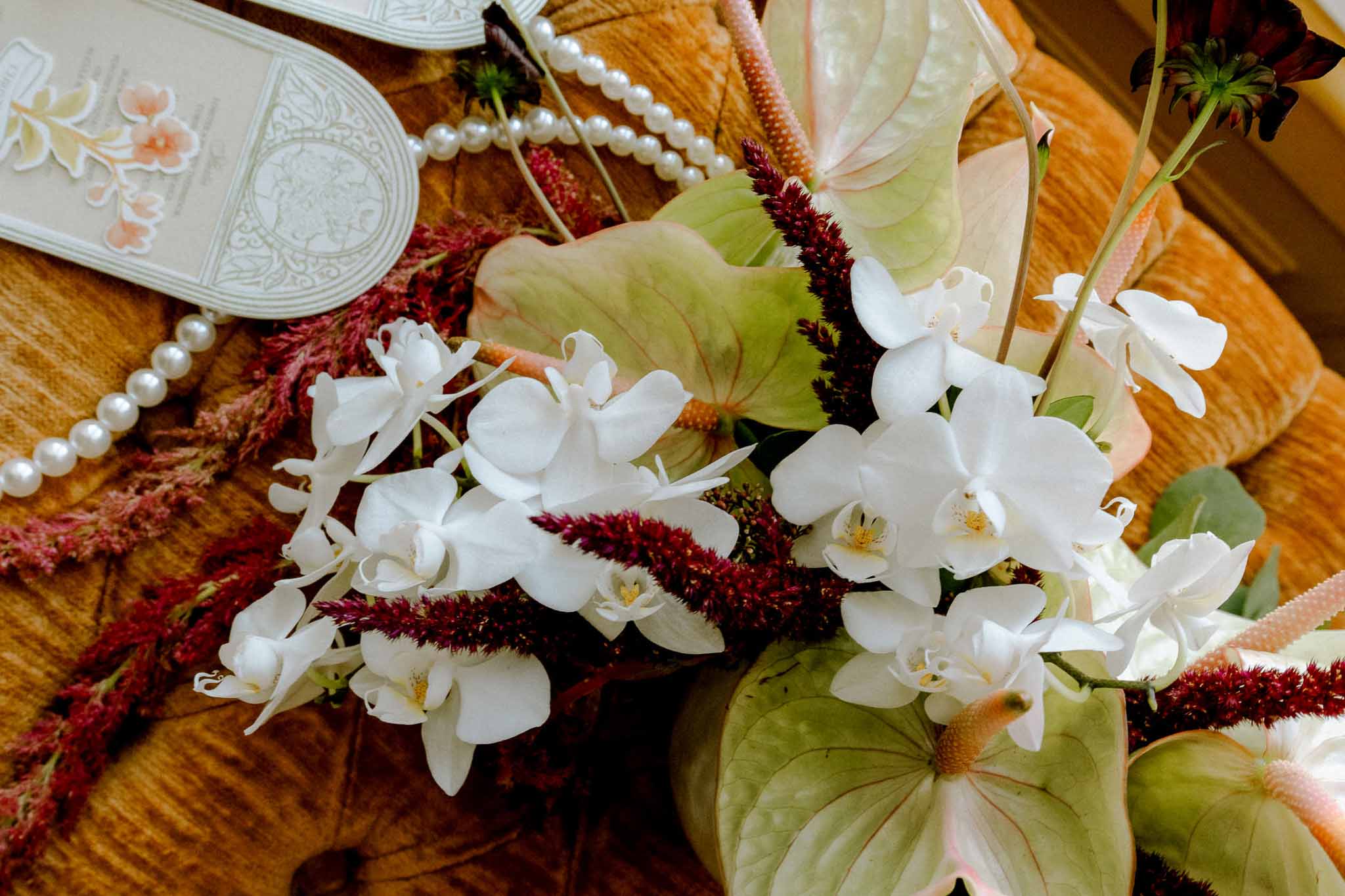 Flat lay of white orchids, burgundy amaranthus, pearl necklace, and illustrated wedding invitation on amber surface