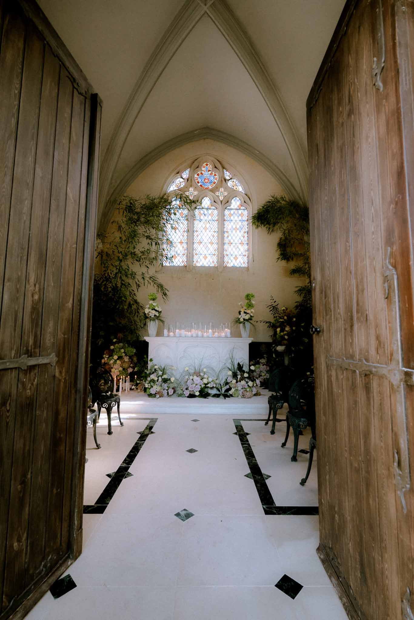 Gothic chapel altar with stained glass, white candles, trailing greenery, and blush ground-level florals