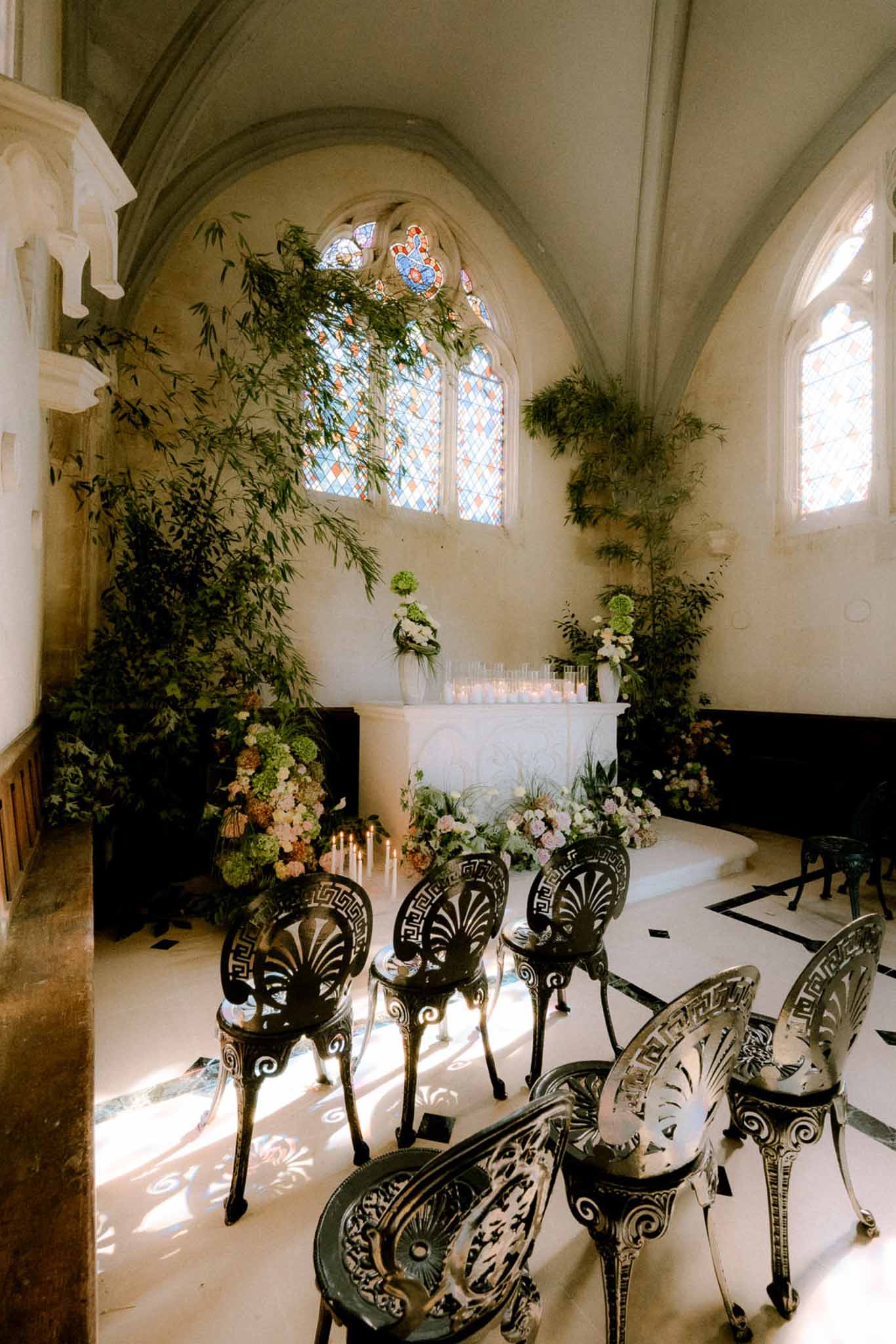 Gothic chapel ceremony setup with stained-glass windows, black iron chairs, and green and blush floral altar arrangements