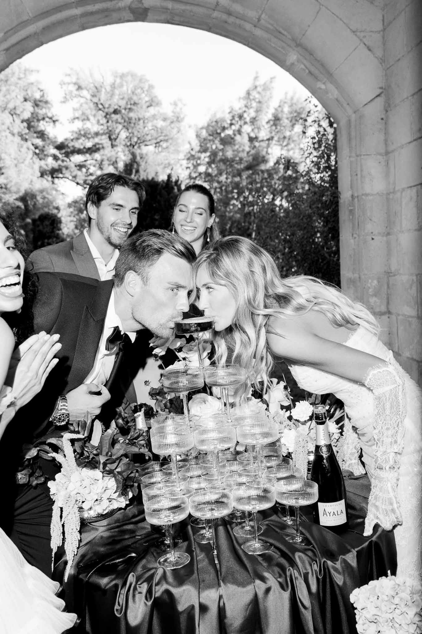 Black and white shot of couple sipping champagne from tower coupe glass beneath stone archway
