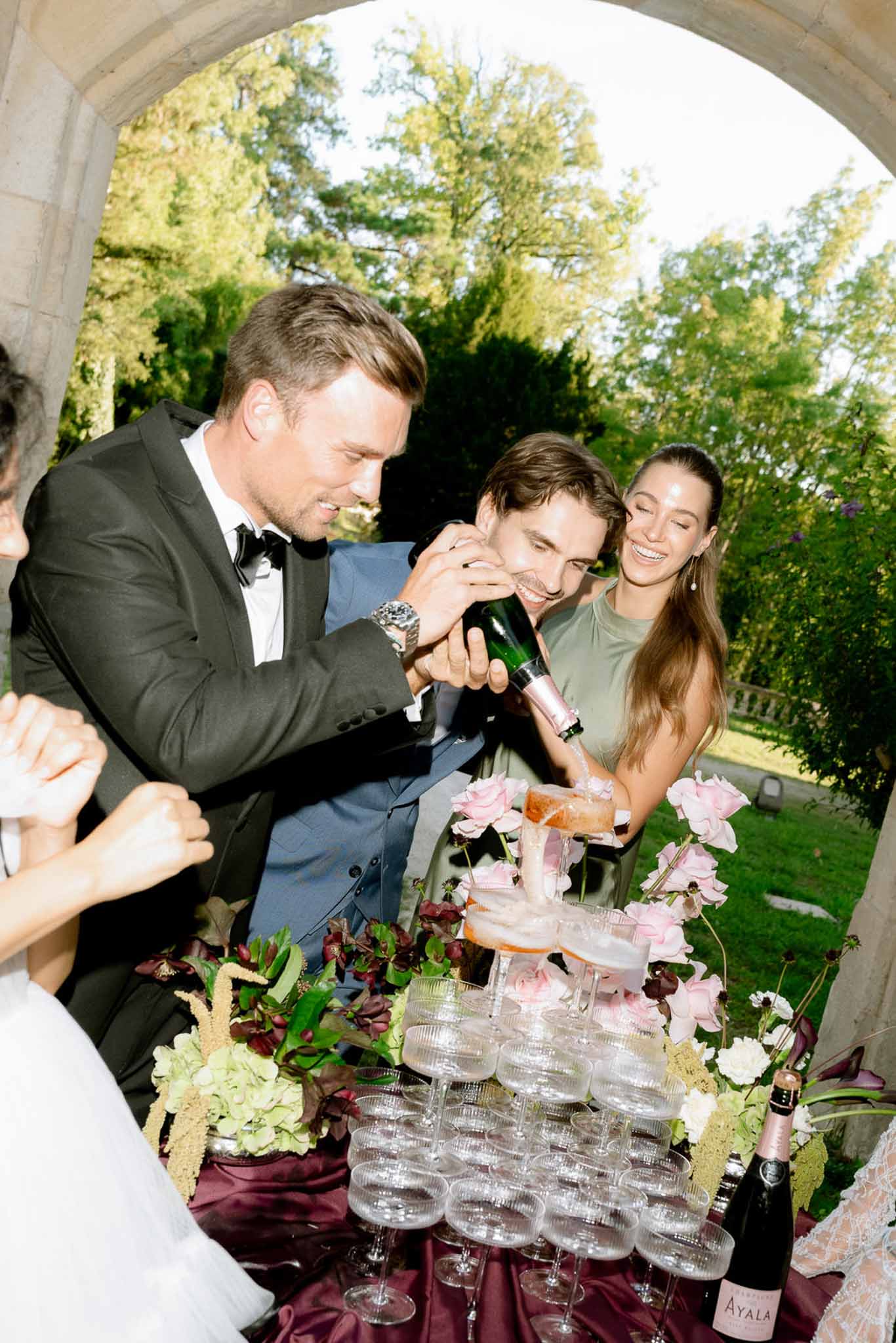 Groom pouring rose champagne over a coupe glass tower beside burgundy floral arrangement at chateau archway