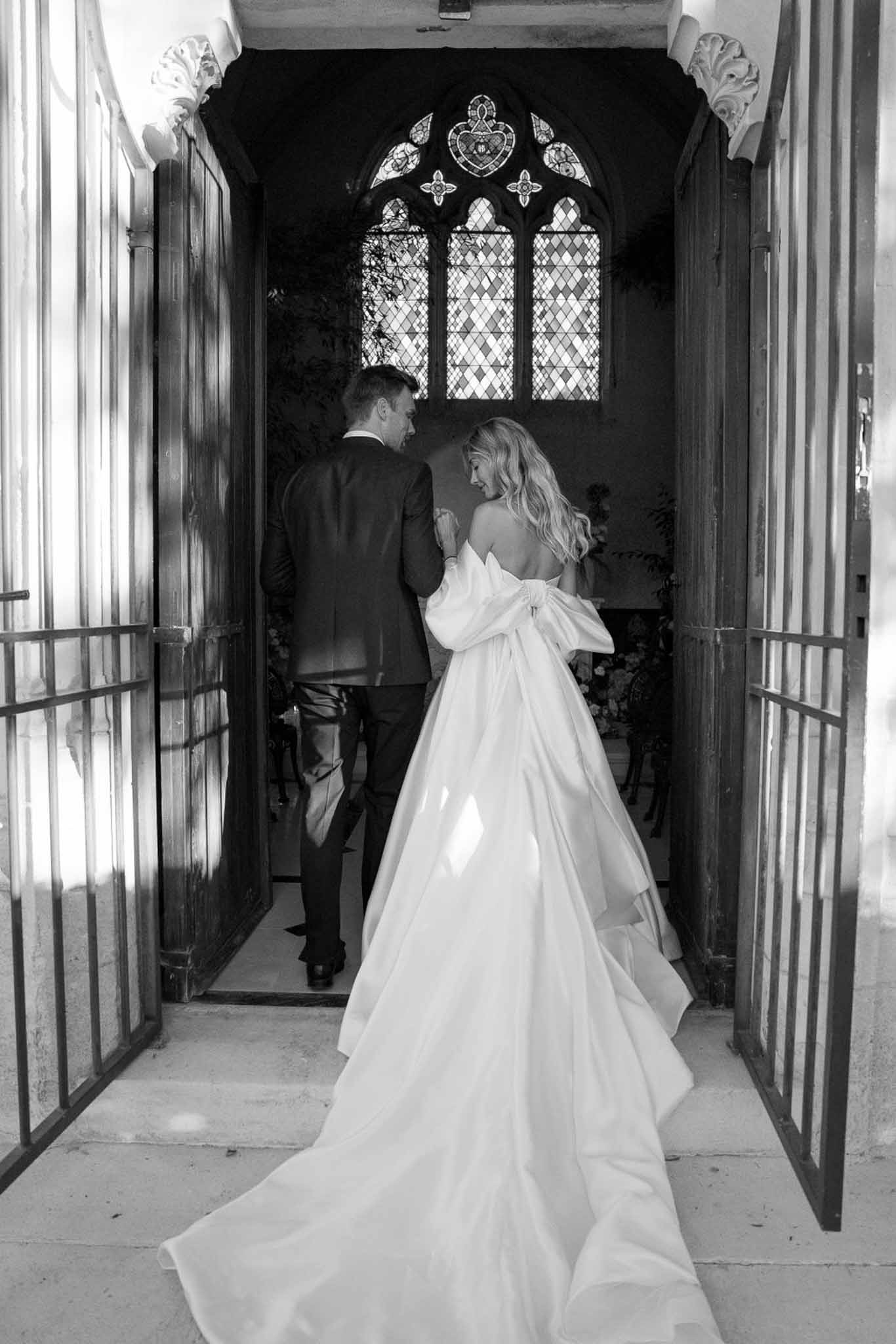 Bride and groom from behind entering chapel through Gothic arch with dramatic train pooling on threshold in B&W