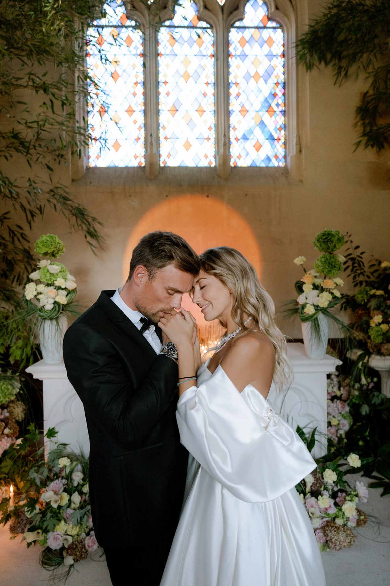 Groom kissing bride's hand at chapel altar with green hydrangea, blush, and cream floral columns and stained glass