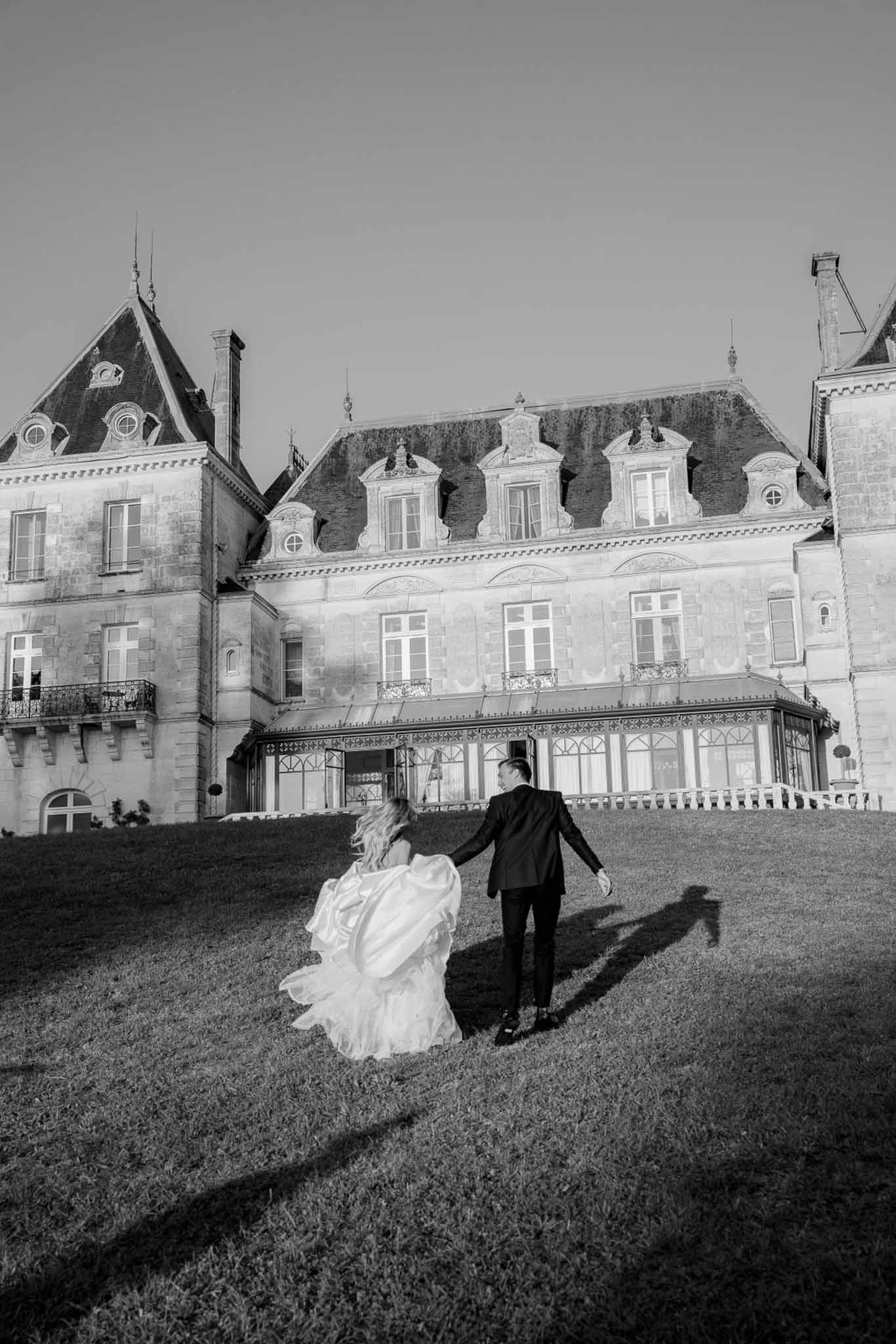 Black and white from behind of couple walking toward mansard-roofed chateau with dramatic train fanning across lawn