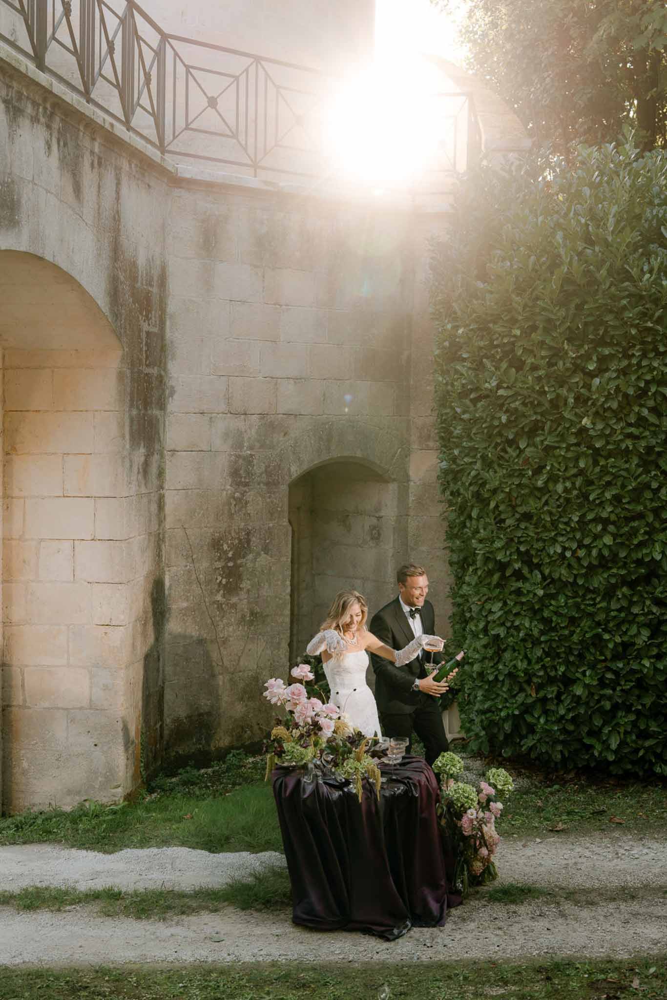 Bride and groom laughing while pouring champagne outside a French chateau with floral arrangement on aubergine linen