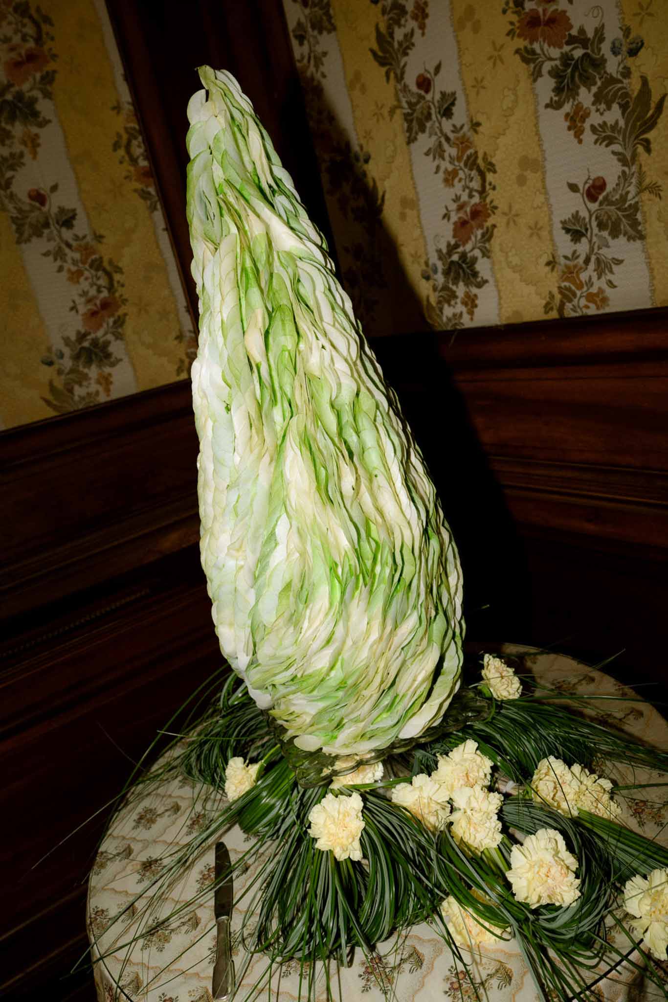 Sculptural floral centerpiece of layered pale green leaves in cone shape with cream carnations on porcelain plate