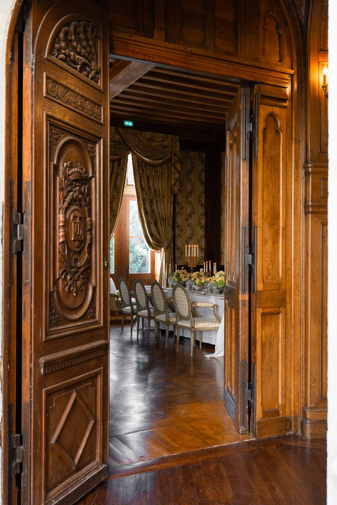 Formal reception dining room viewed through carved double doors with long banquet table, gold candelabra, and sage green c...