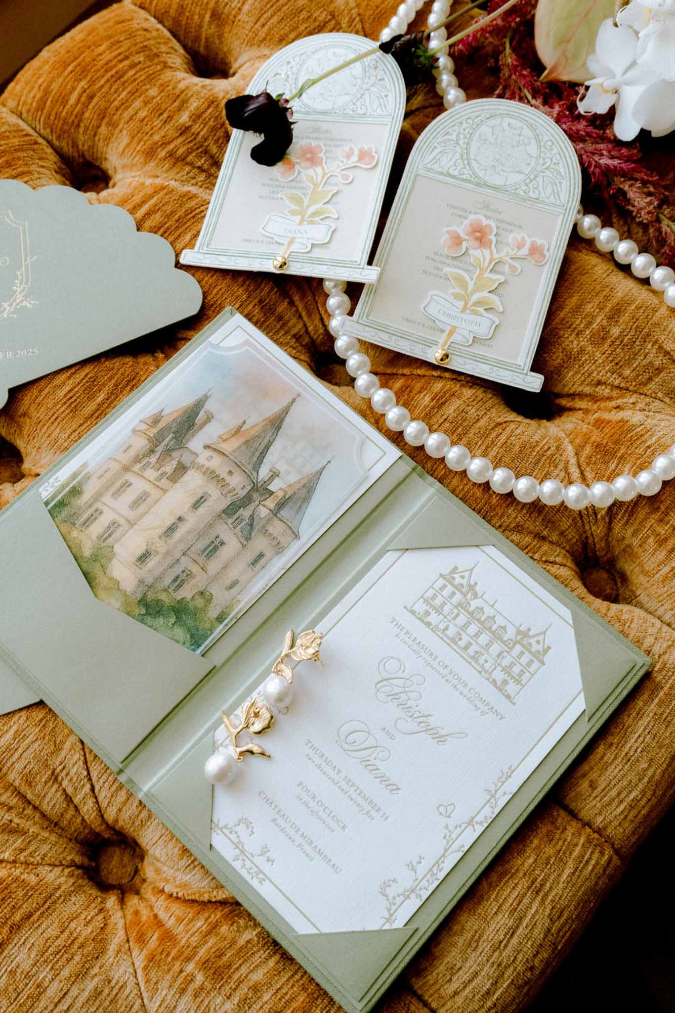Flat lay of sage green and cream wedding stationery suite with gold script, pearl necklace, and floral charm on velvet sur...