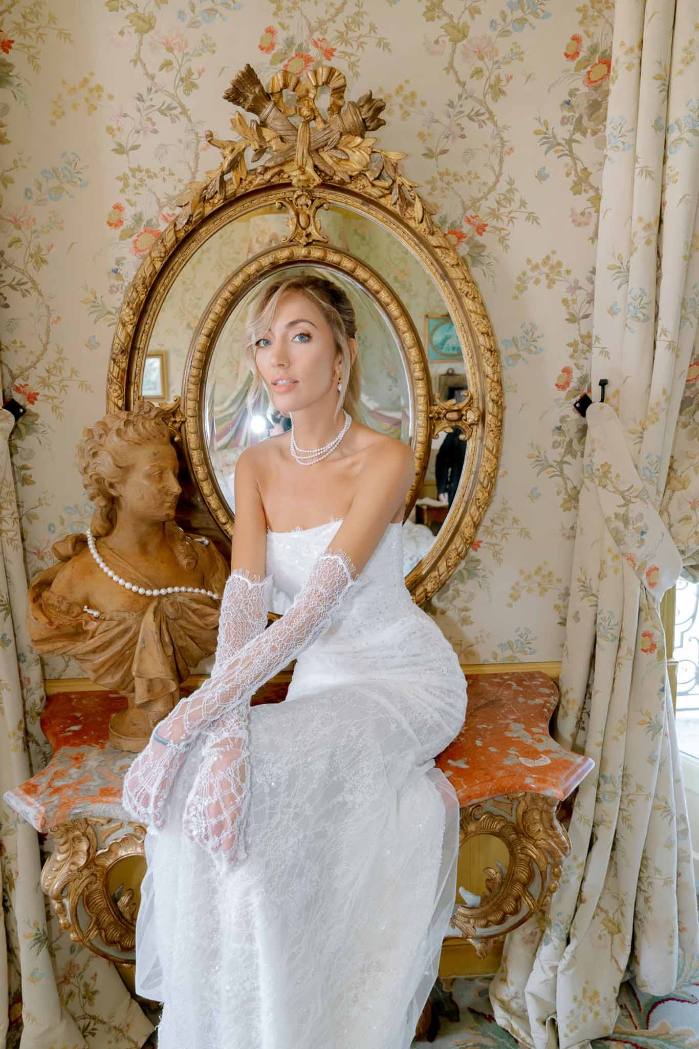 Bride in strapless lace gown and pearl necklace seated on console table in chateau dressing room with gilt mirror