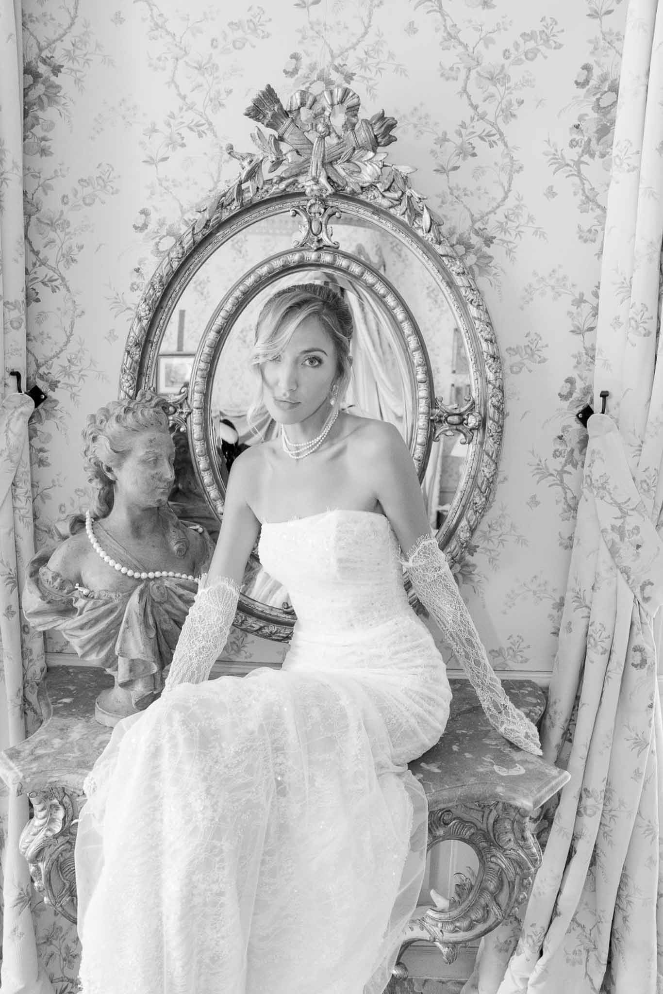 Black-and-white portrait of bride in strapless lace gown seated before ornate mirror in chateau dressing room