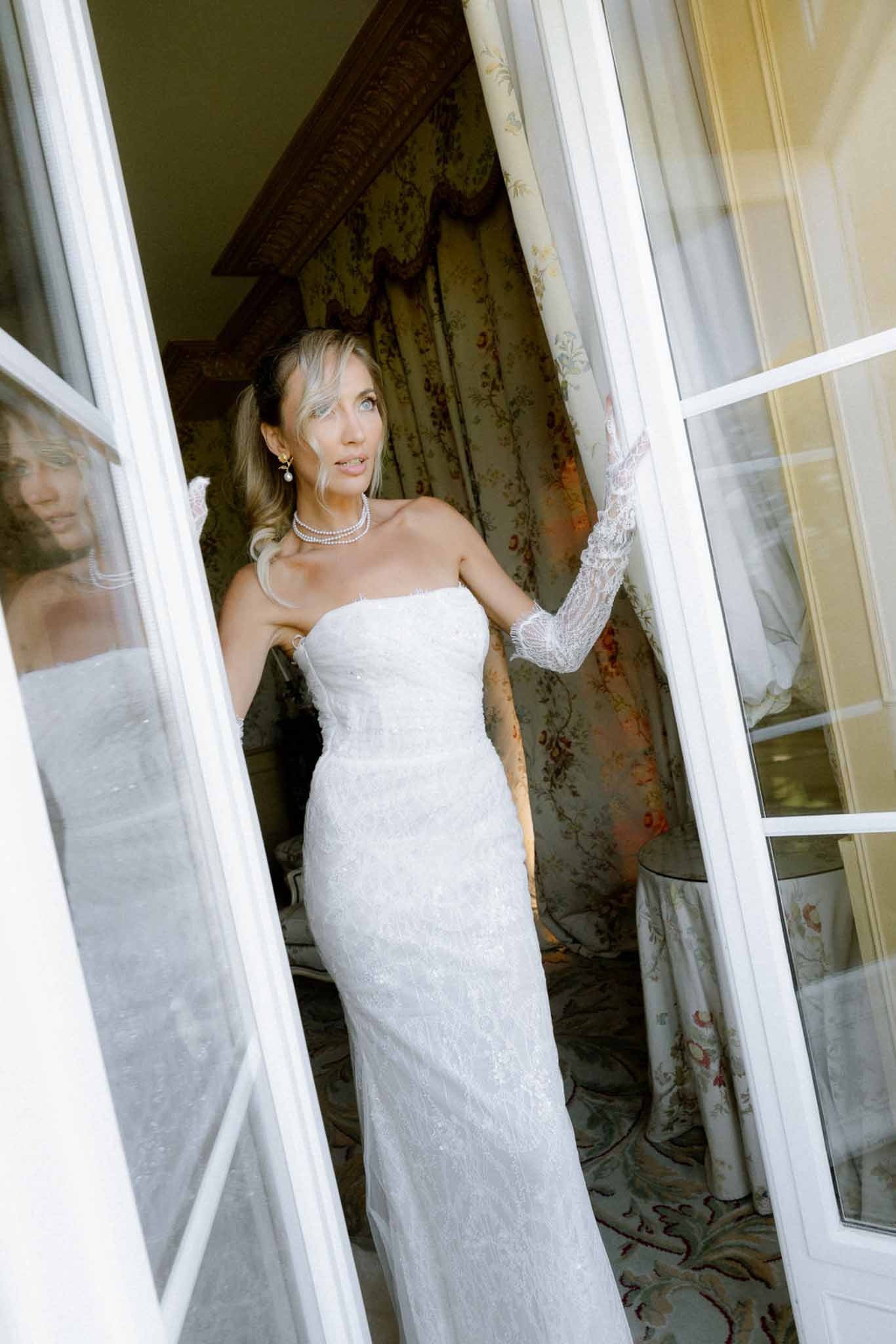 Bride in strapless white lace gown with pearl choker standing in French doors of chateau during getting ready
