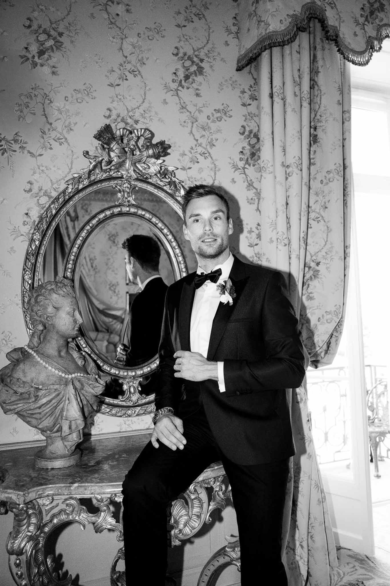 Black and white portrait of groom in tuxedo leaning against gilt console beside carved mirror