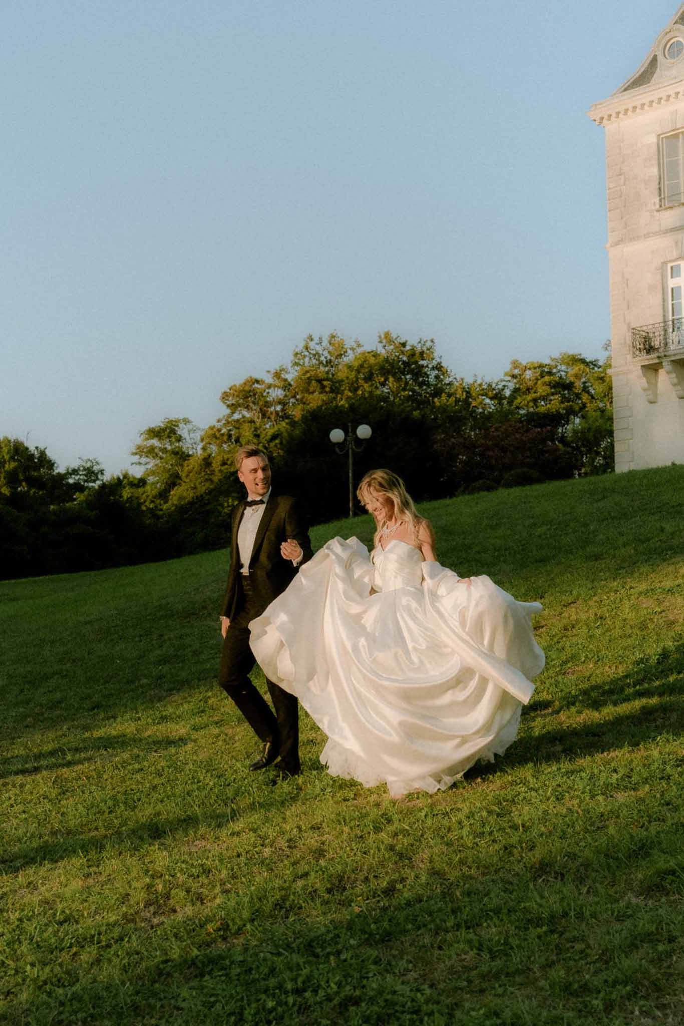 Bride in ivory ballgown and groom in black tuxedo walking on chateau lawn at golden hour