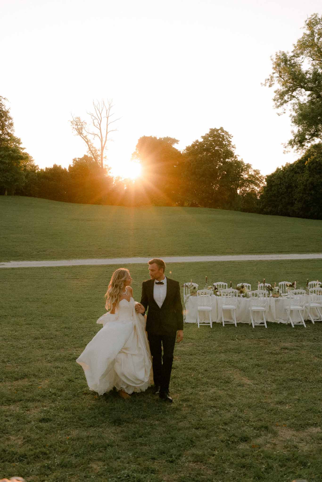 Couple walking hand-in-hand across lawn at golden hour with dressed reception table visible in background