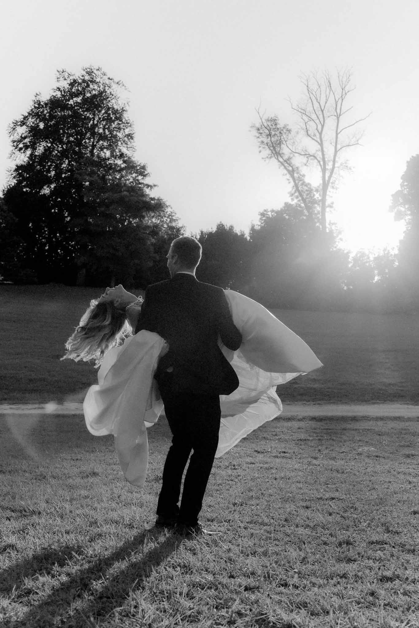 Black-and-white backlit shot of groom carrying bride across an open field with flowing cape billowing