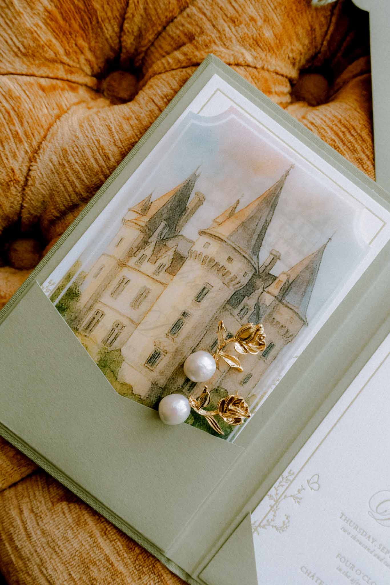 Wedding invitation in sage envelope with chateau illustration, pearl and gold earrings on velvet surface