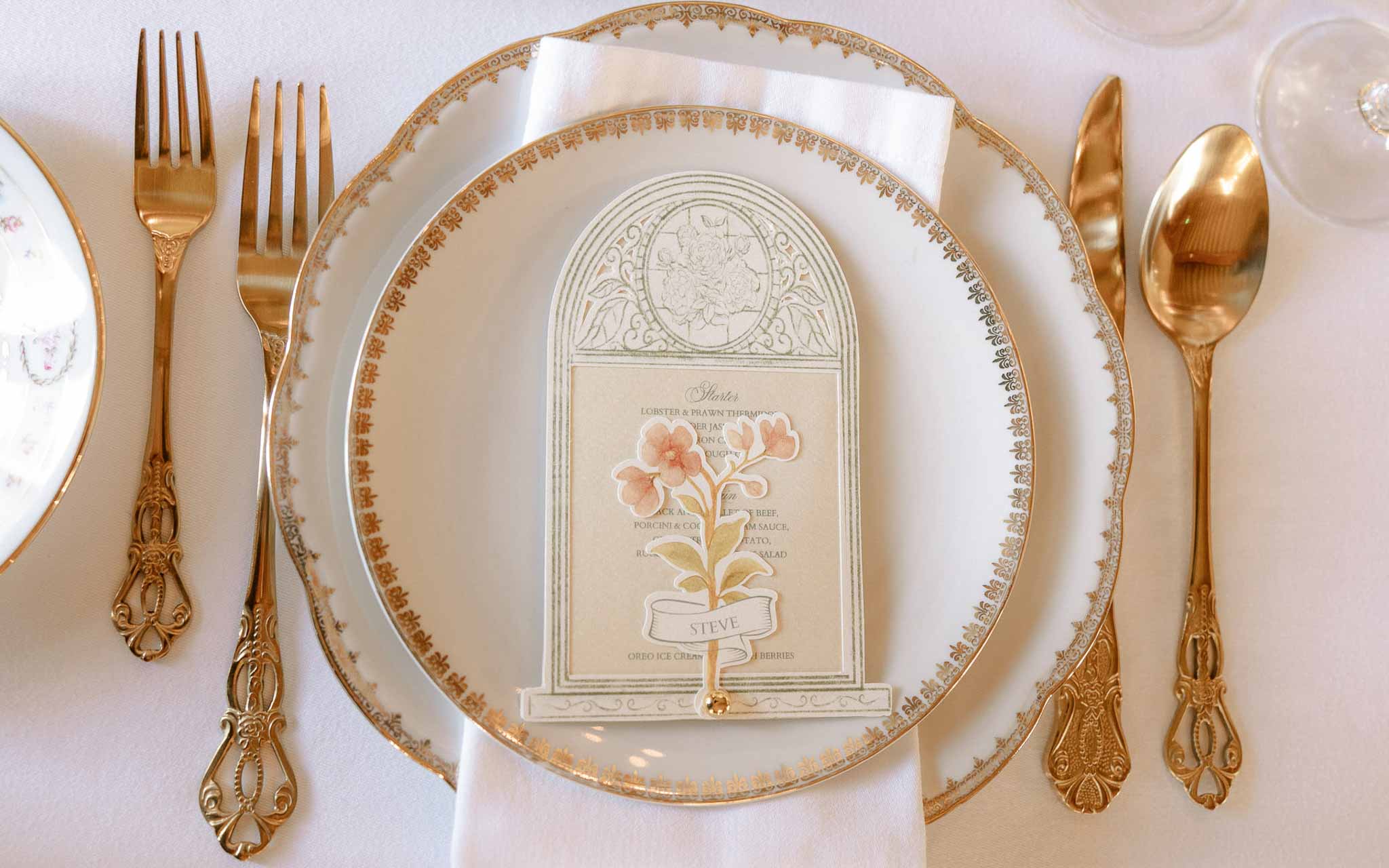 Gold-rimmed charger with botanical arch menu card in sage and blush, ornate gold cutlery on white linen
