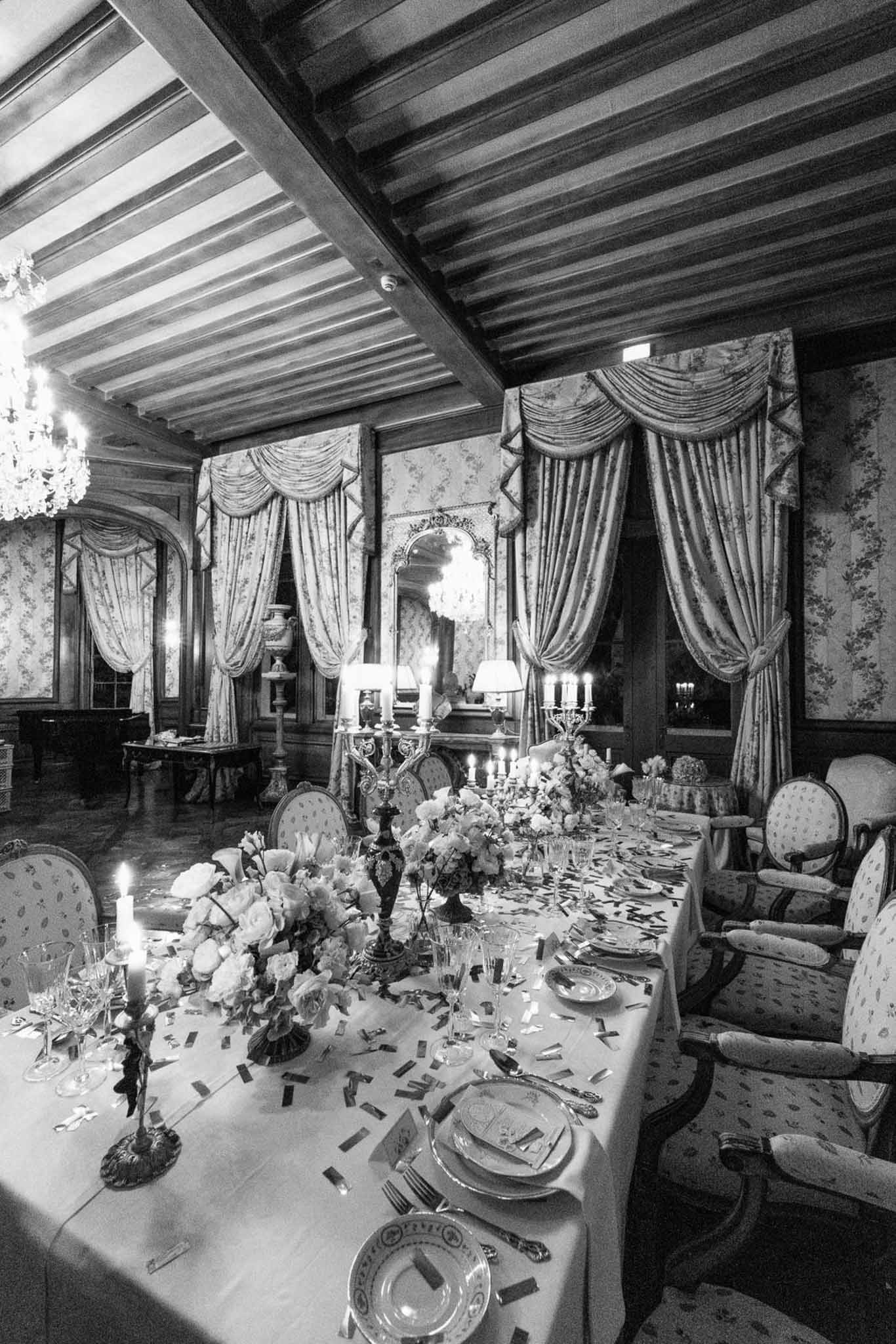 Black and white wide shot of a long reception dinner table with candelabras and floral centerpieces in a chateau salon