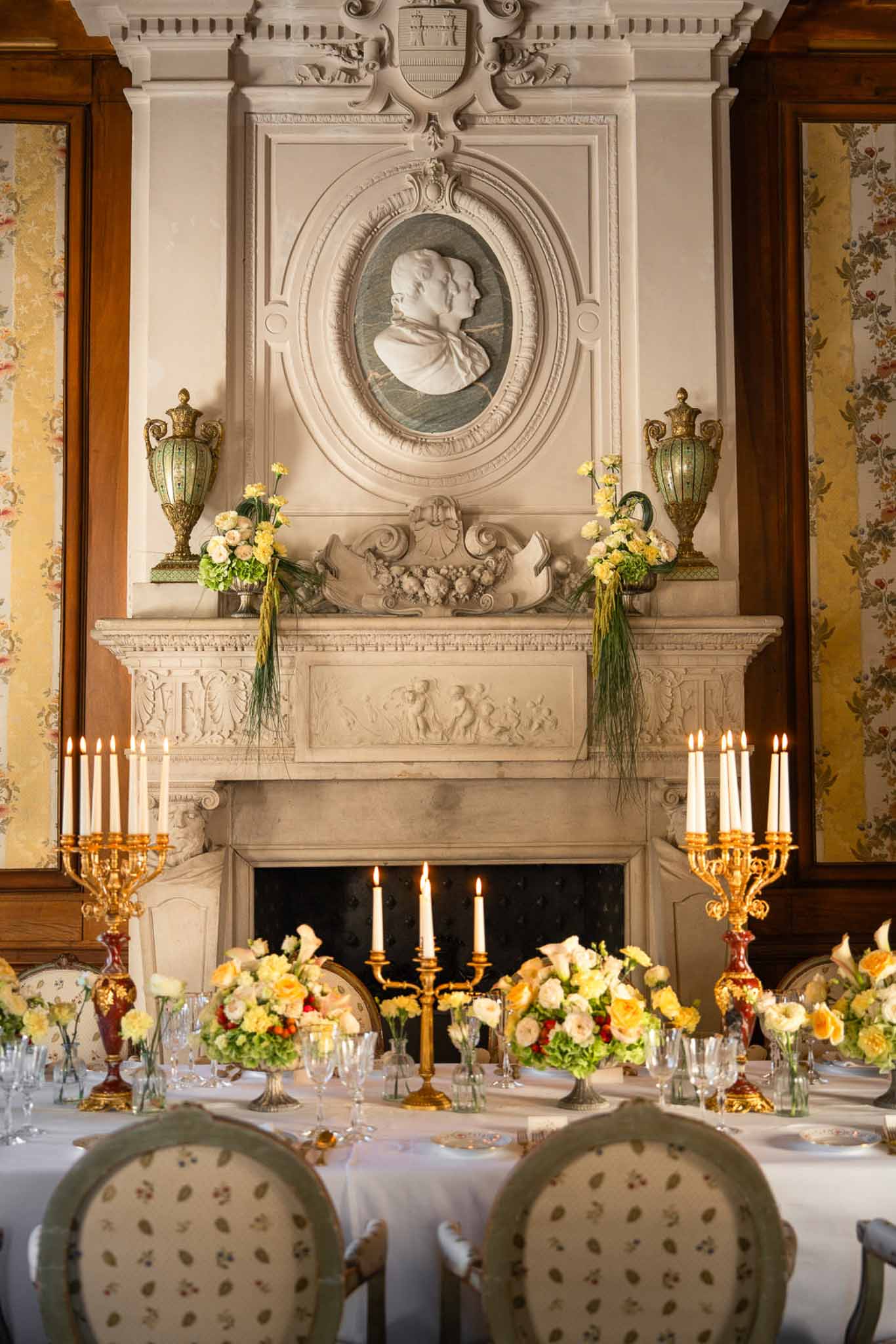 Yellow and peach centerpieces with gold candelabras before carved fireplace in floral-papered dining room