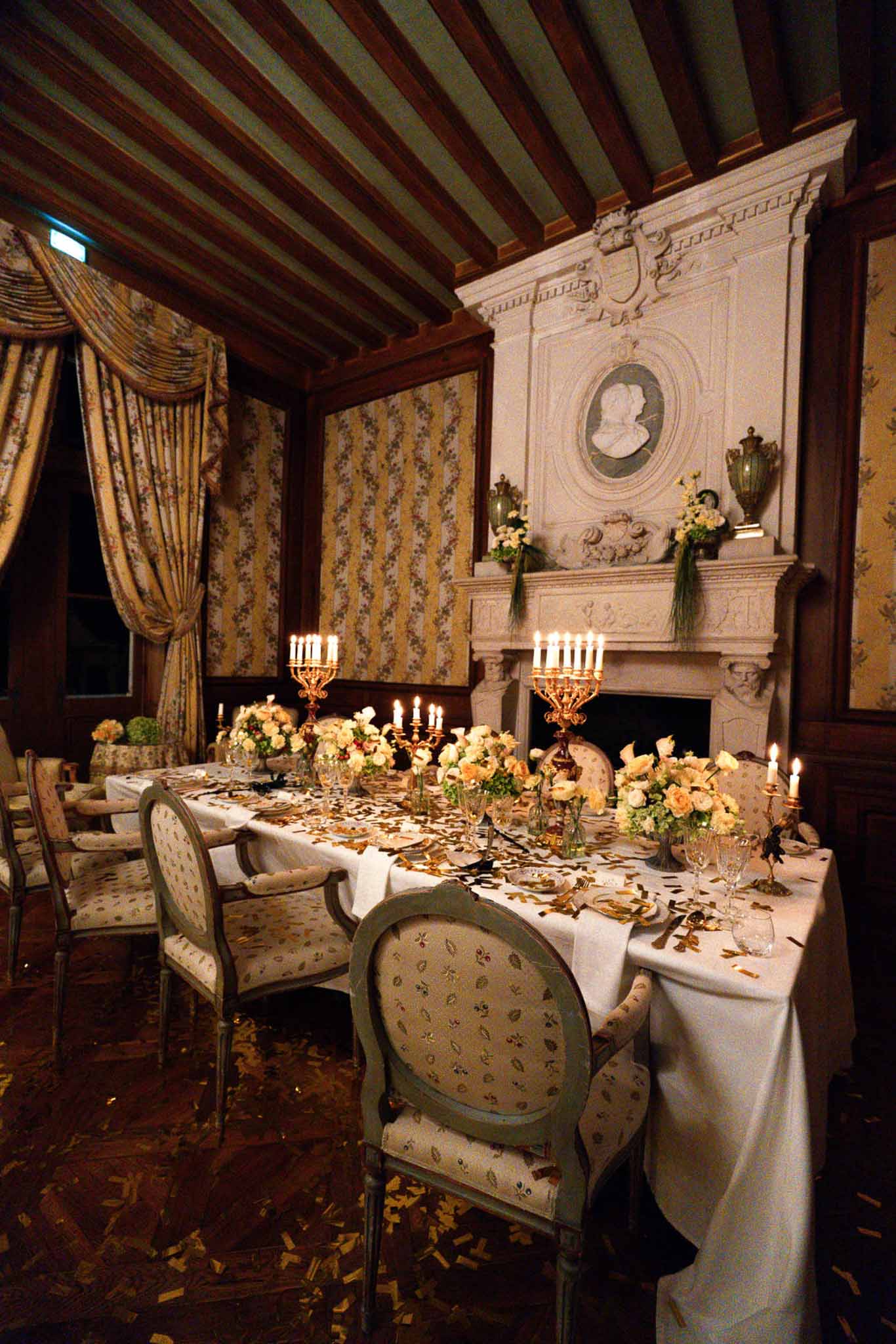 Gold candelabras with peach roses on long table before carved fireplace in floral-wallpapered chateau salon