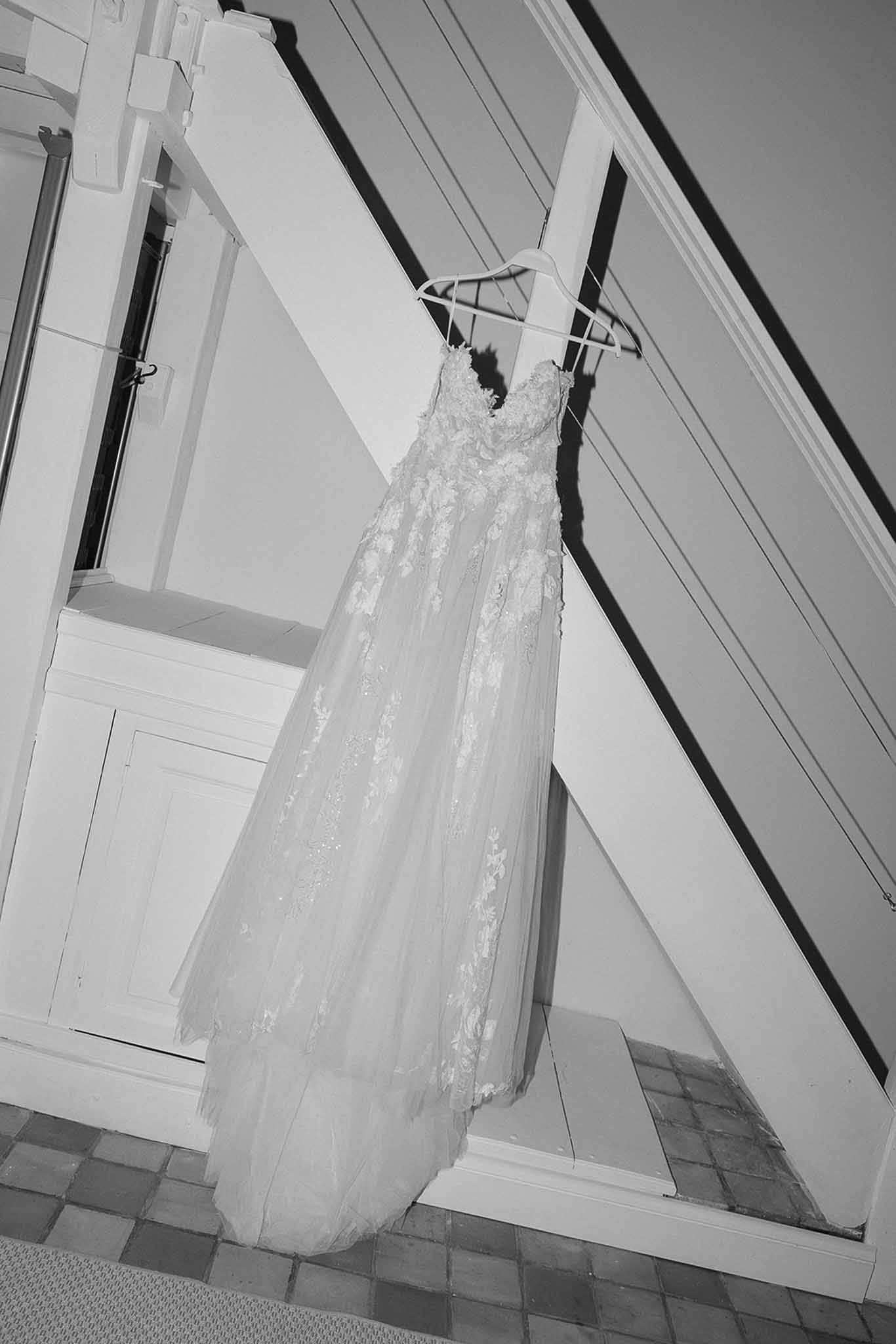 Black-and-white photo of a lace-appliqued wedding dress with tulle skirt hanging on a staircase banister