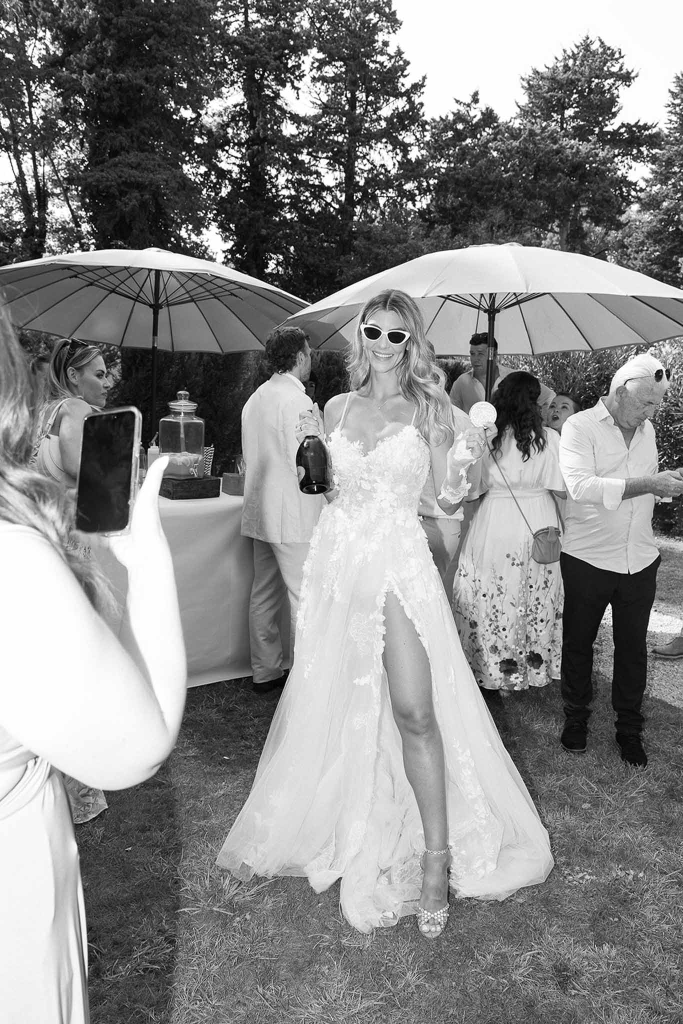 Black and white candid of bride in floral tulle gown holding champagne bottle with cat-eye sunglasses