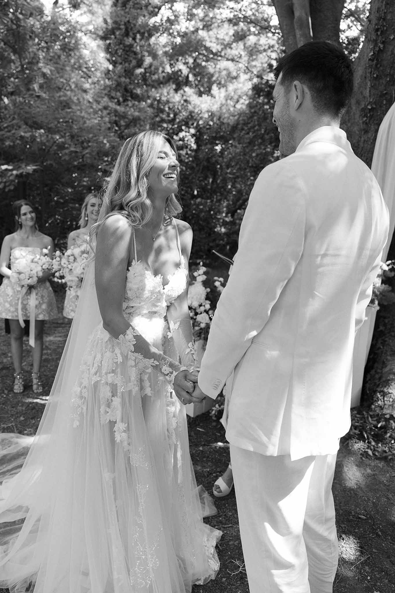 Bride and groom holding hands during outdoor ceremony, bride laughing in floral applique gown, groom in white suit