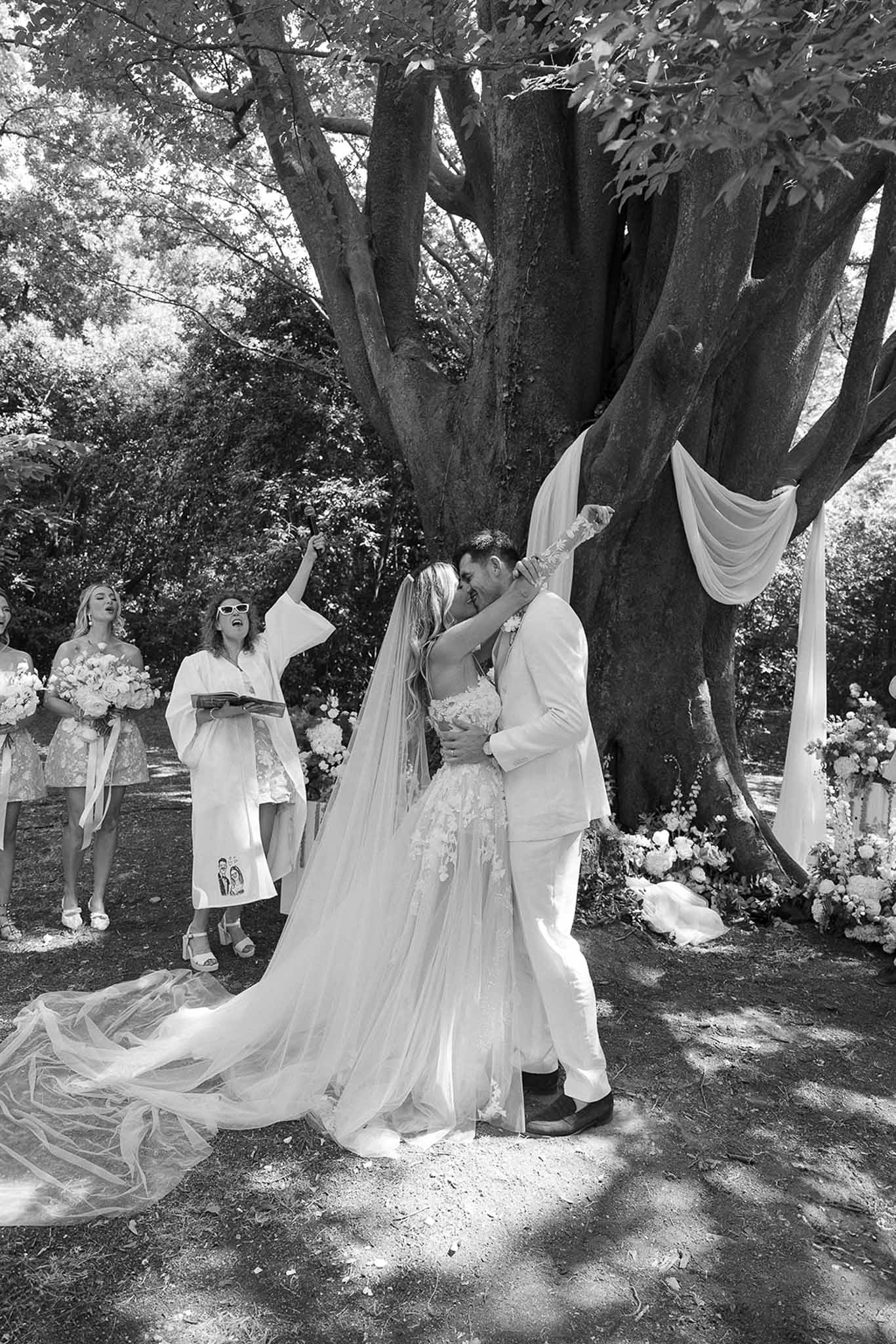 Black and white photo of bride and groom sharing first kiss under a decorated tree during outdoor ceremony