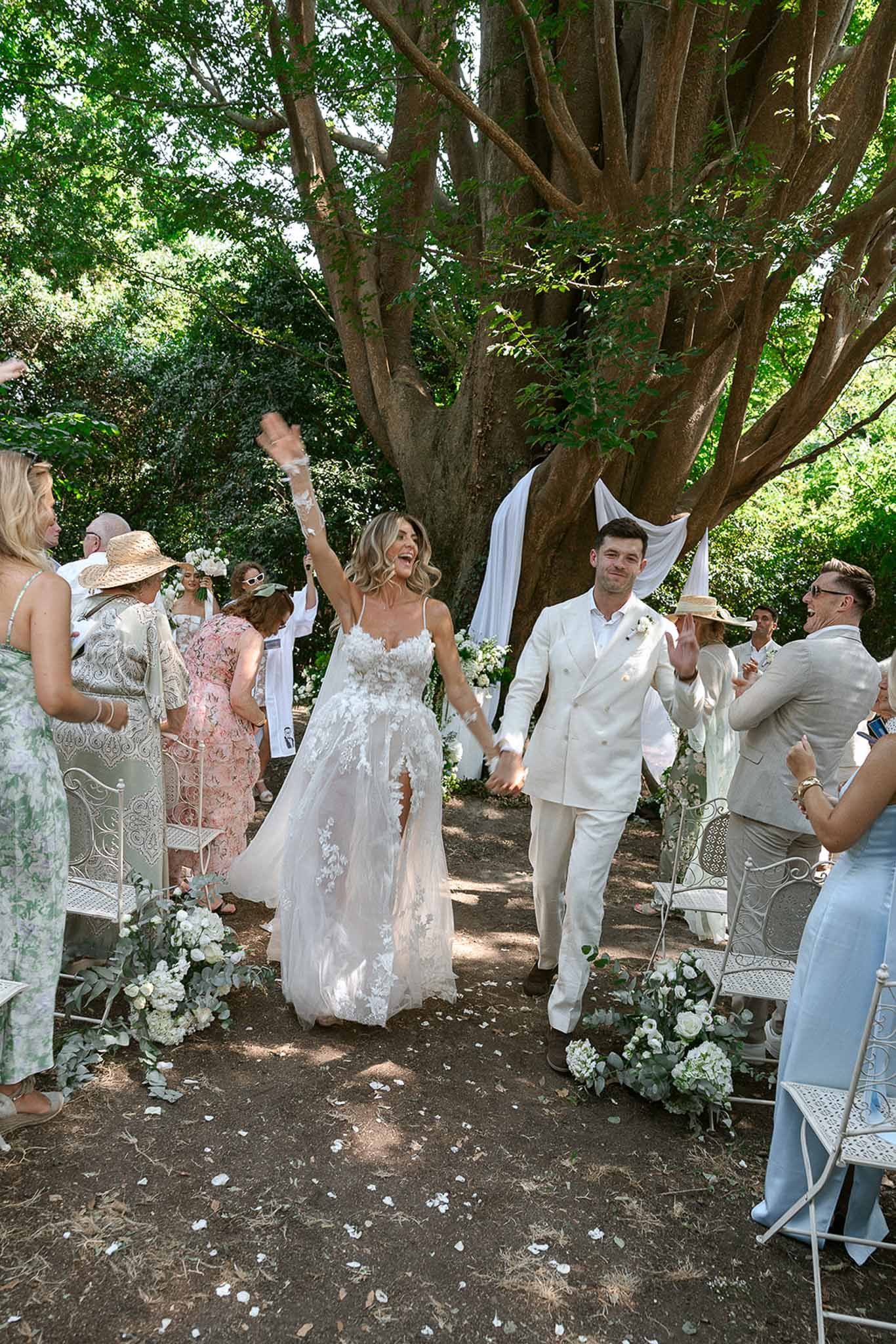 Couple walking back up garden aisle with bride's arm raised in celebration under draped tree