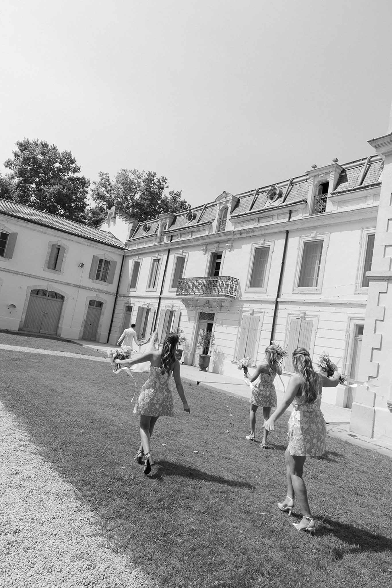 Black and white rear shot of bride and bridesmaids running across lawn toward mansard-roofed chateau