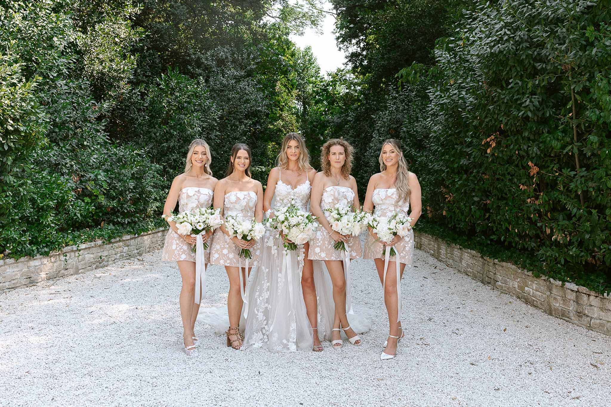 Bride and four bridesmaids in blush floral mini dresses holding white rose bouquets on gravel path