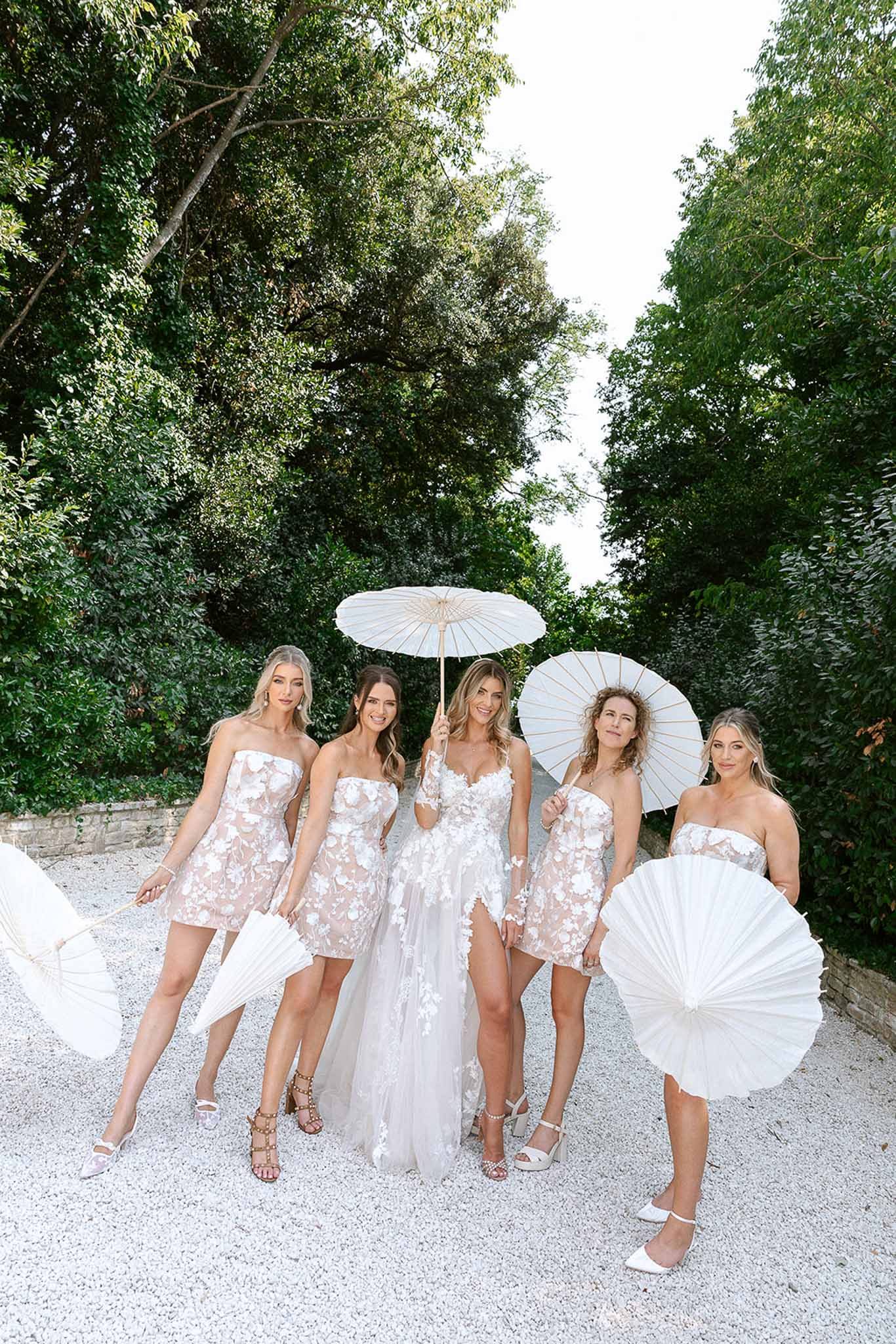 Bride and four bridesmaids in coordinating white floral dresses holding paper parasols on a gravel path