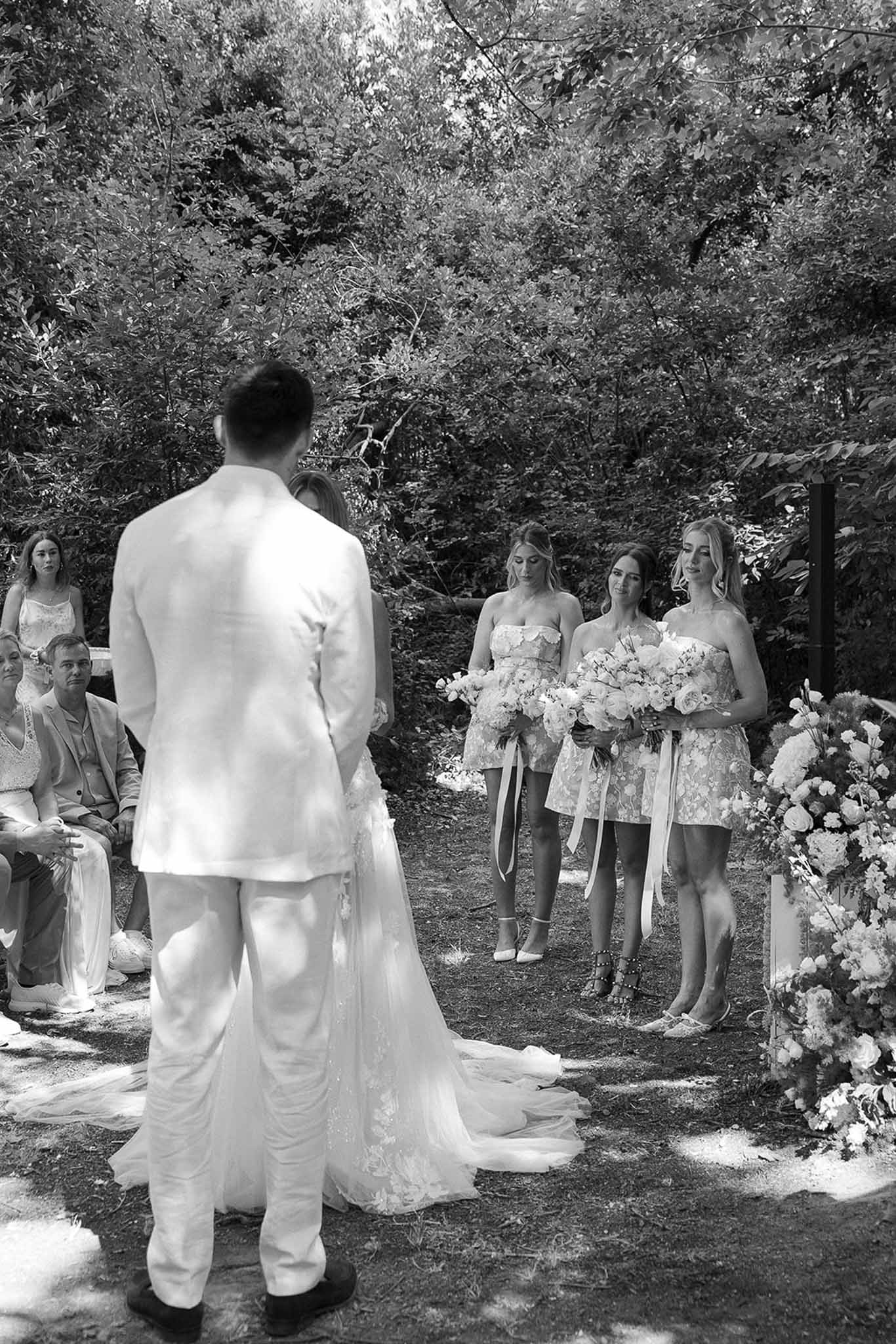 Black and white from behind of couple at altar with three bridesmaids in floral dresses holding trailing bouquets