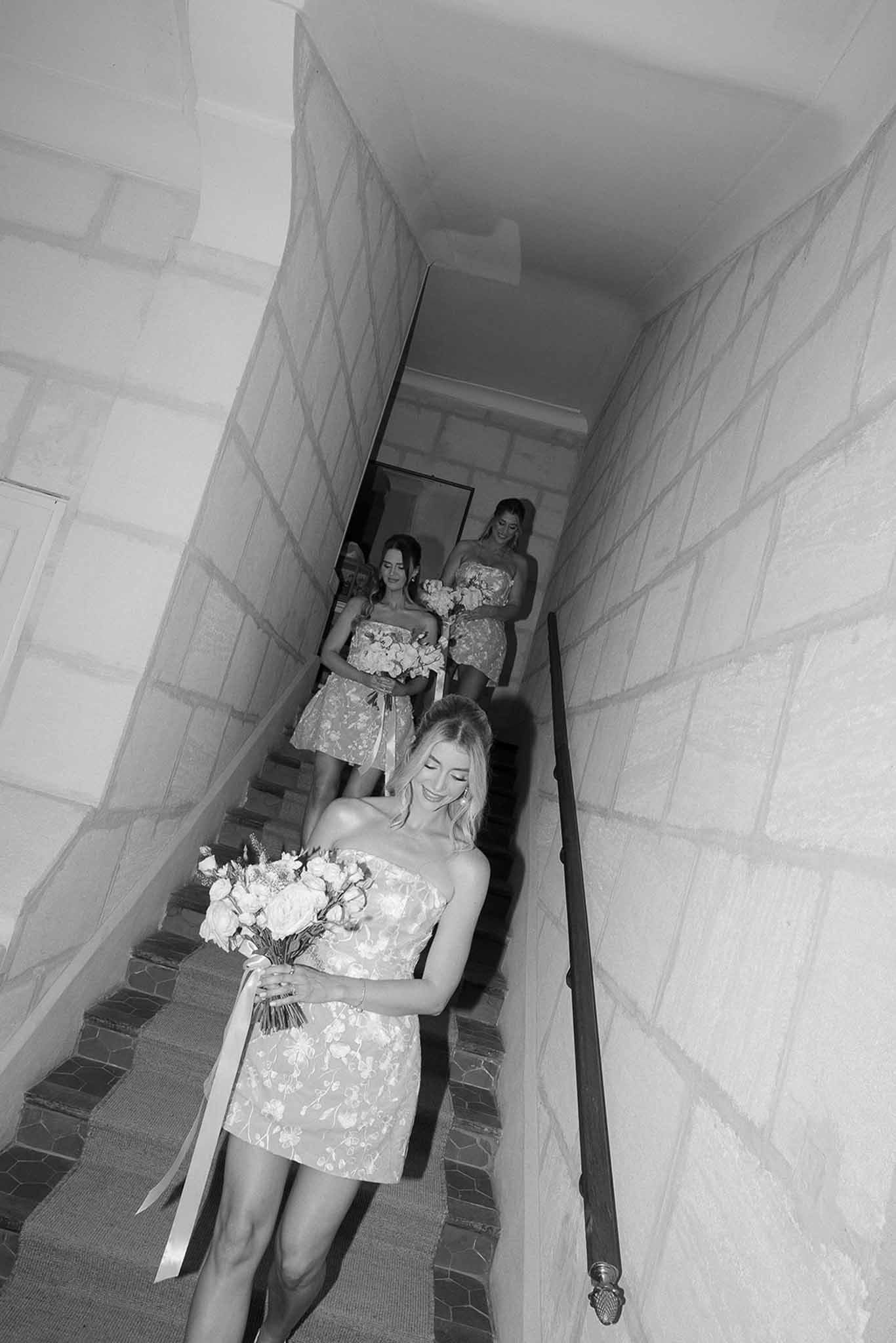 Black and white three bridesmaids in strapless floral dresses descending stone staircase carrying bouquets