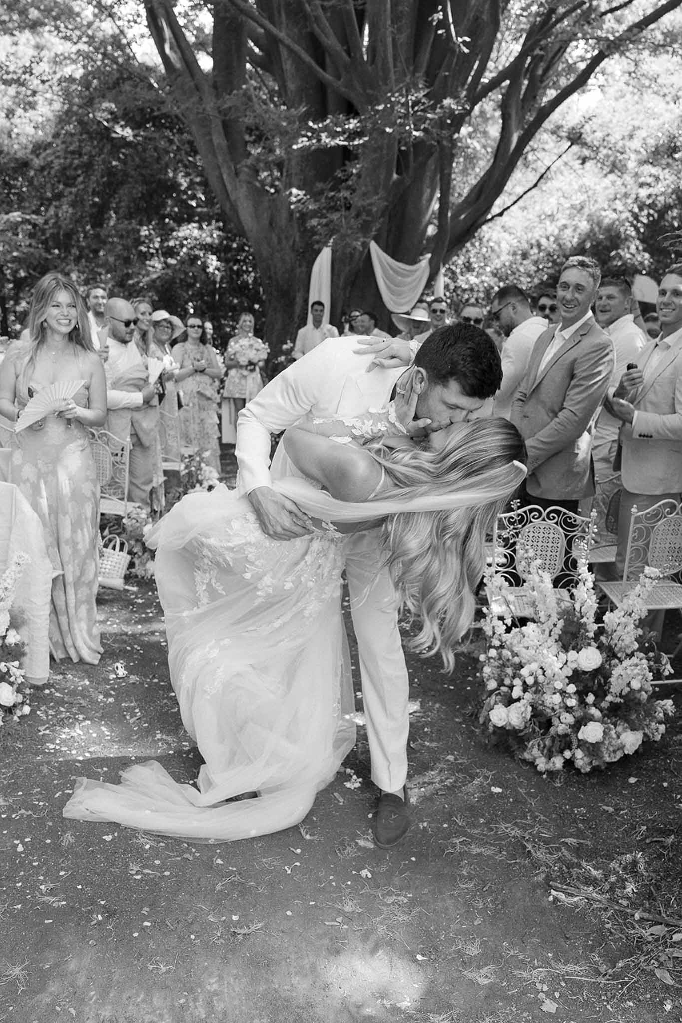 Black and white photo of groom dipping bride for first kiss as guests applaud in outdoor ceremony aisle