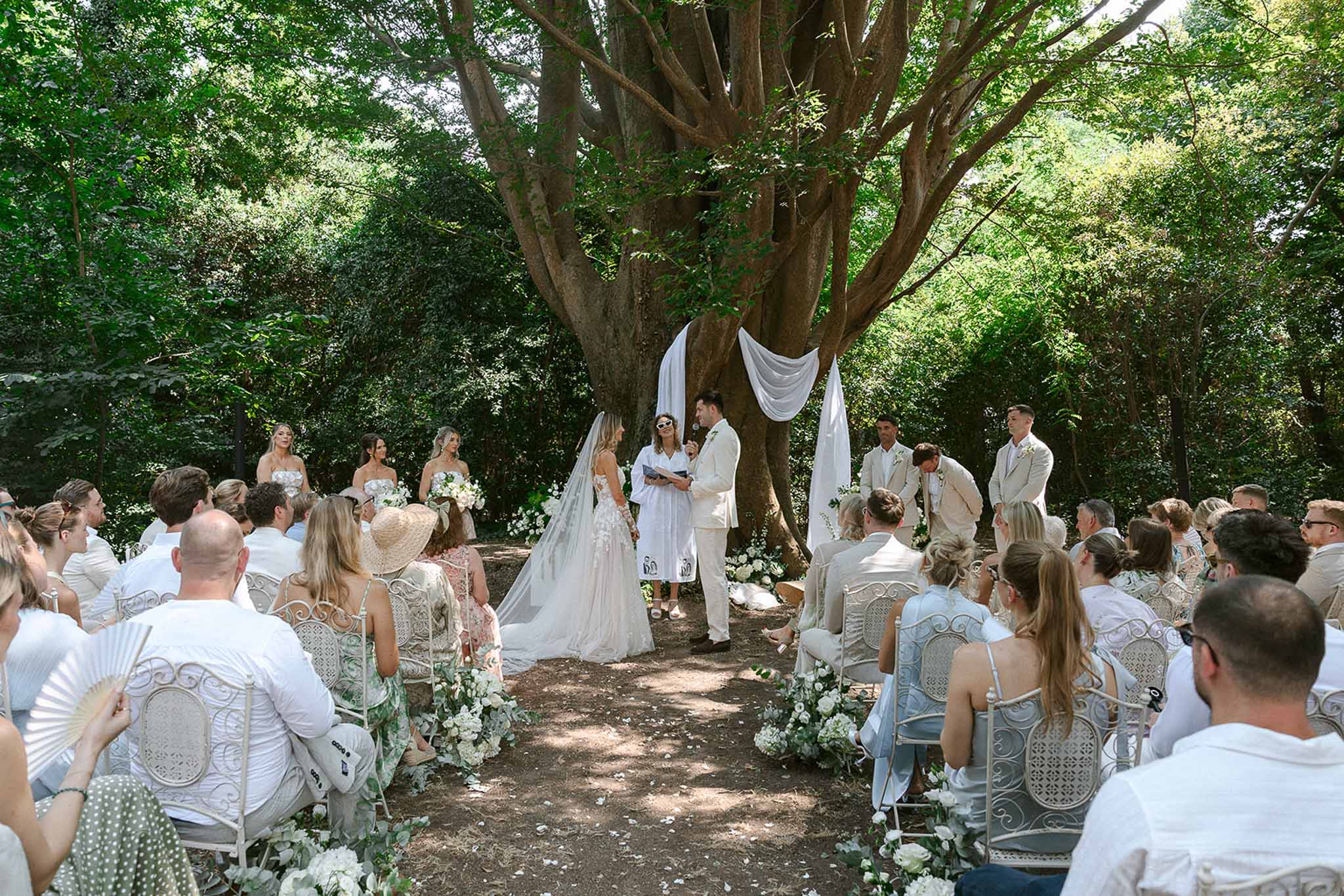 All-white ceremony under tree with draped fabric bridesmaids in ivory print groomsmen in cream sixty guests on iron chairs