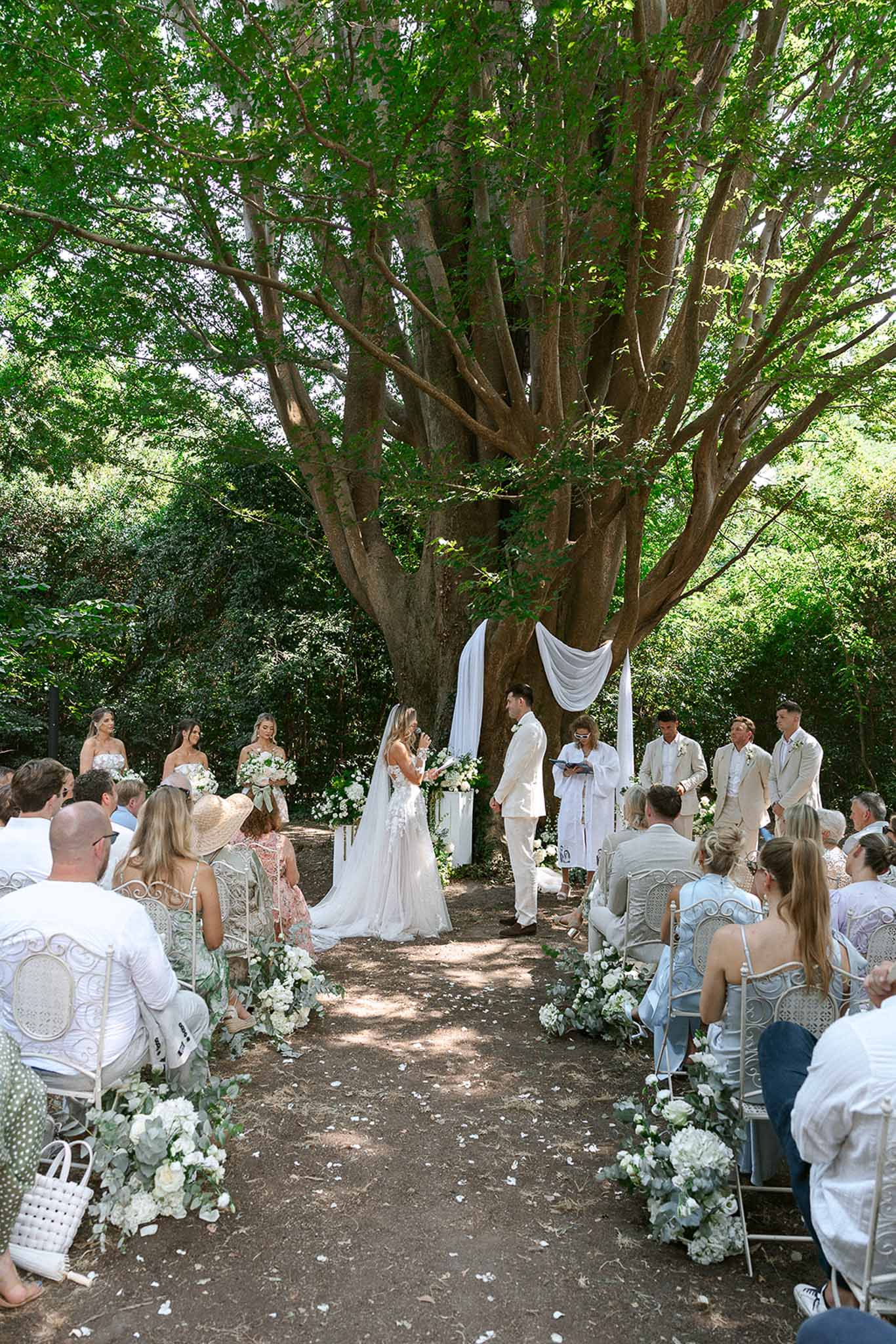 Garden ceremony under spreading tree with white floral aisle arrangements and bridal party in white