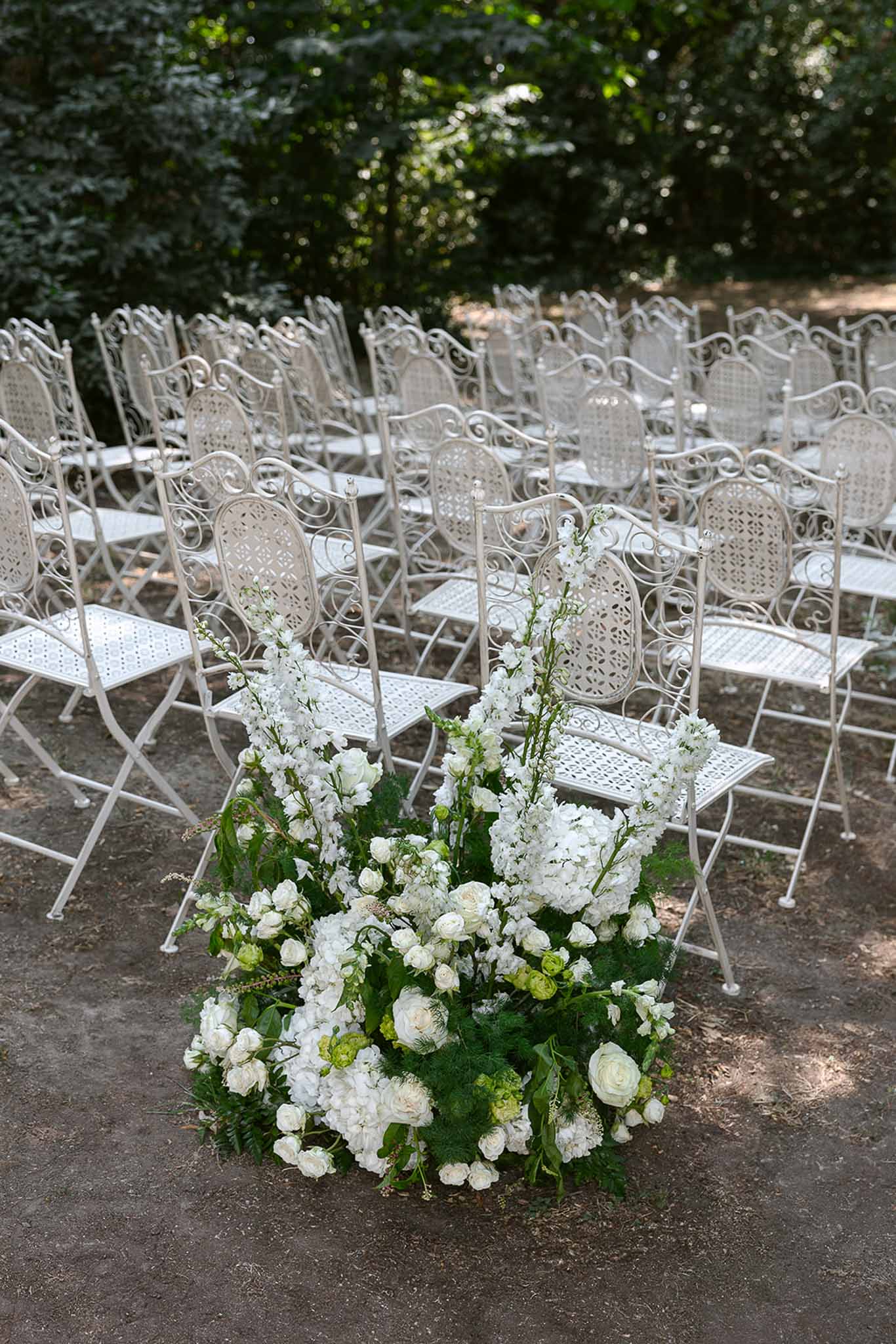 Ceremony aisle with white rose hydrangea and fern ground arrangement before wrought-iron chairs