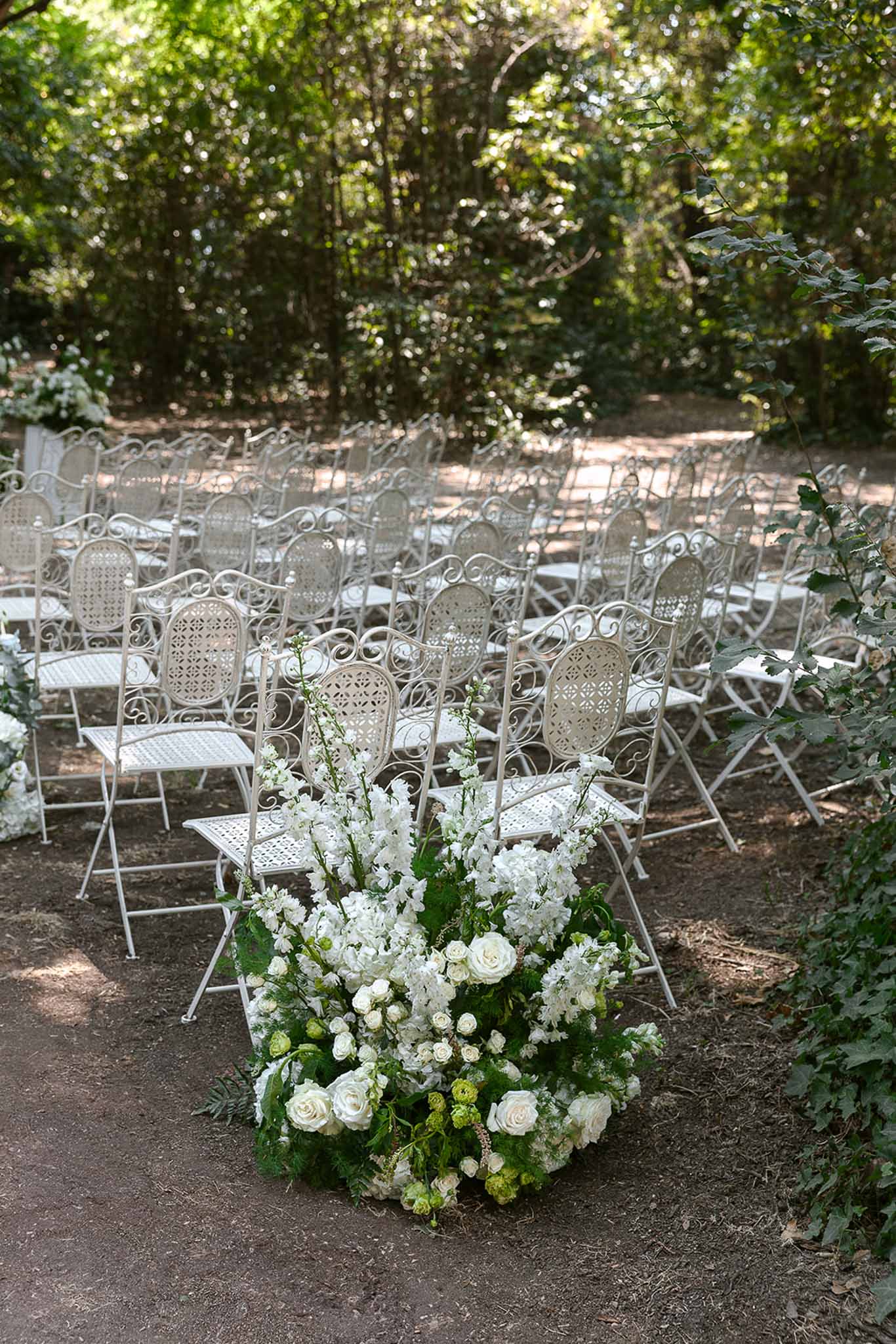 Cream wrought-iron bistro chairs flanking aisle with white rose and hydrangea ground arrangement in wooded garden