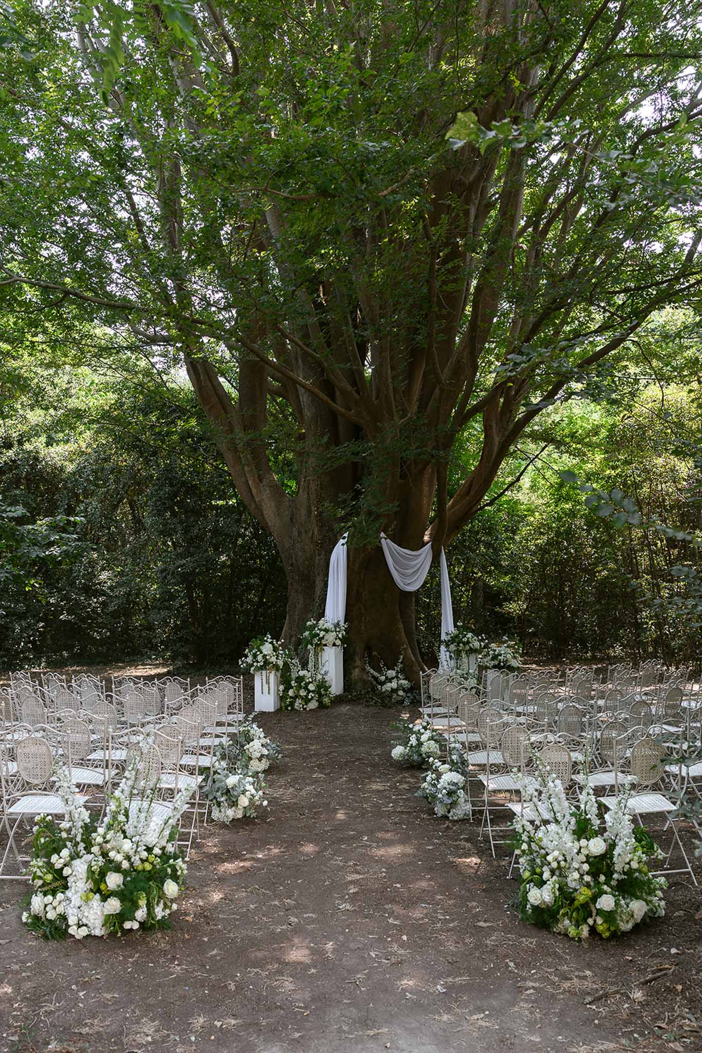 Outdoor ceremony setup with white chairs, floral aisle arrangements, and draped fabric altar beneath a large tree