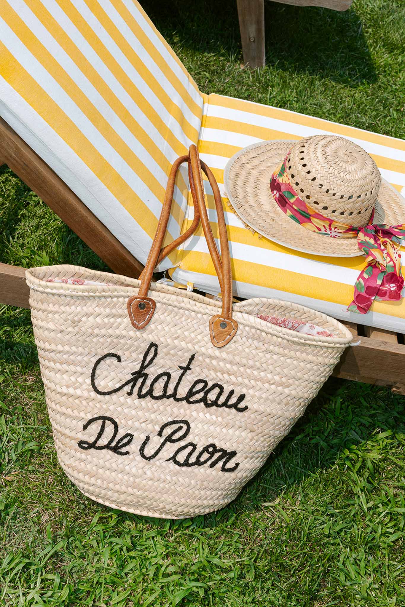 Straw tote bag embroidered with Chateau De Paon and sun hat on striped yellow lounger poolside