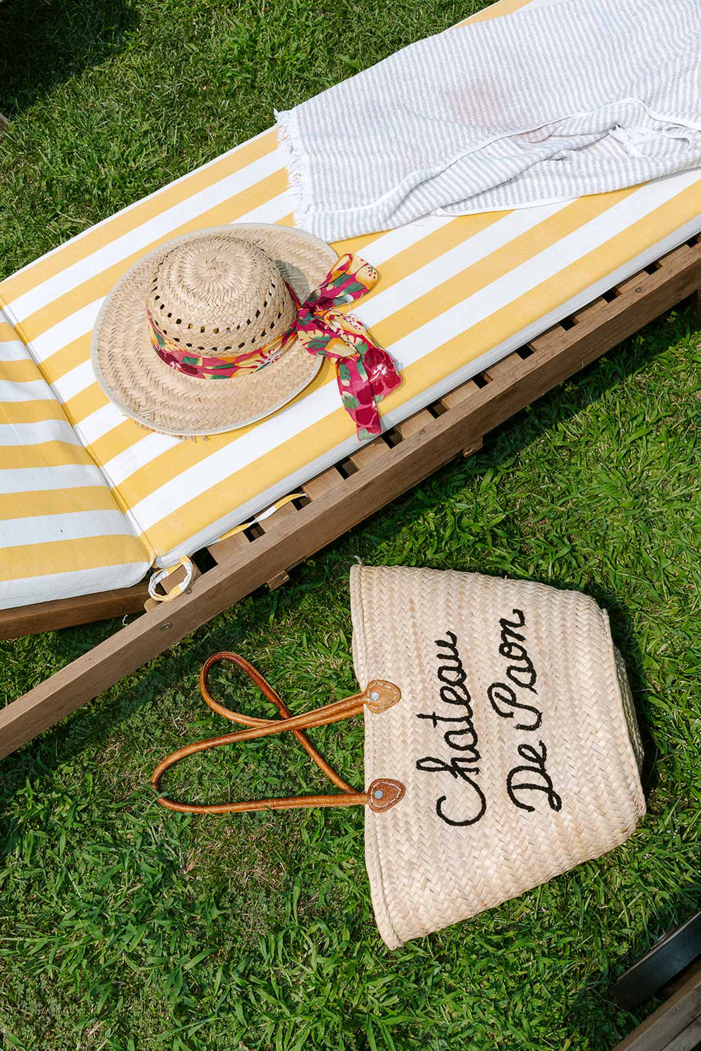 Yellow striped sun lounger with straw hat, towel, and branded Chateau De Paon tote bag on grass lawn