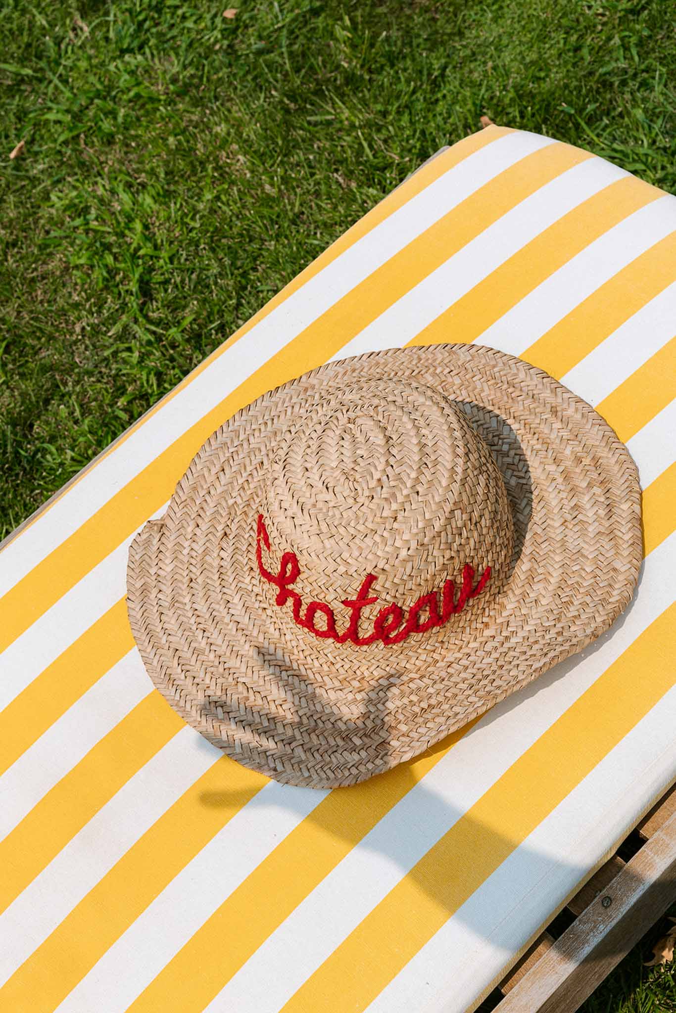 Straw hat embroidered with chateau in red cursive placed on a yellow striped sun lounger cushion