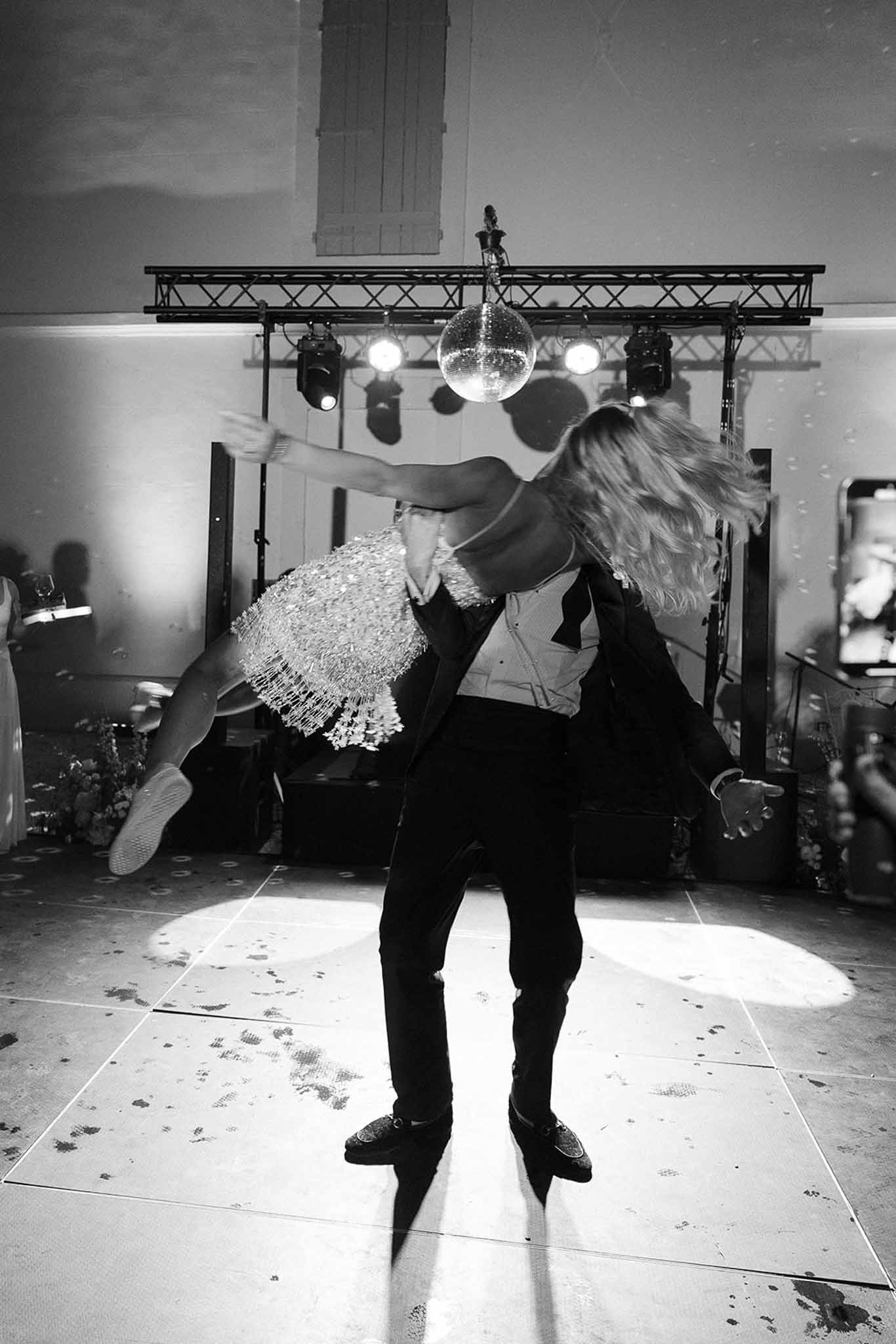 Man lifting woman in embellished mini dress under disco ball with stage spotlights on dance floor in B&W