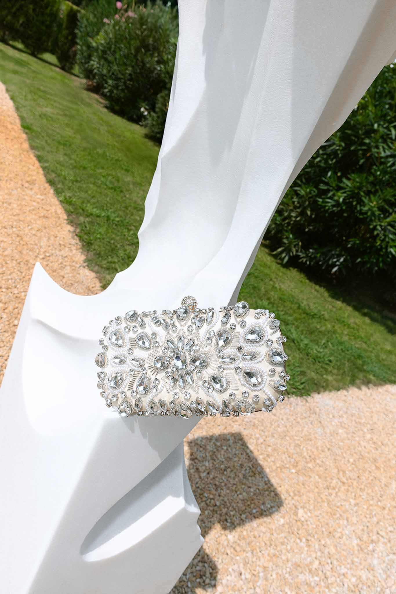 Crystal-encrusted ivory bridal clutch bag with rhinestone floral pattern on white draped surface in garden