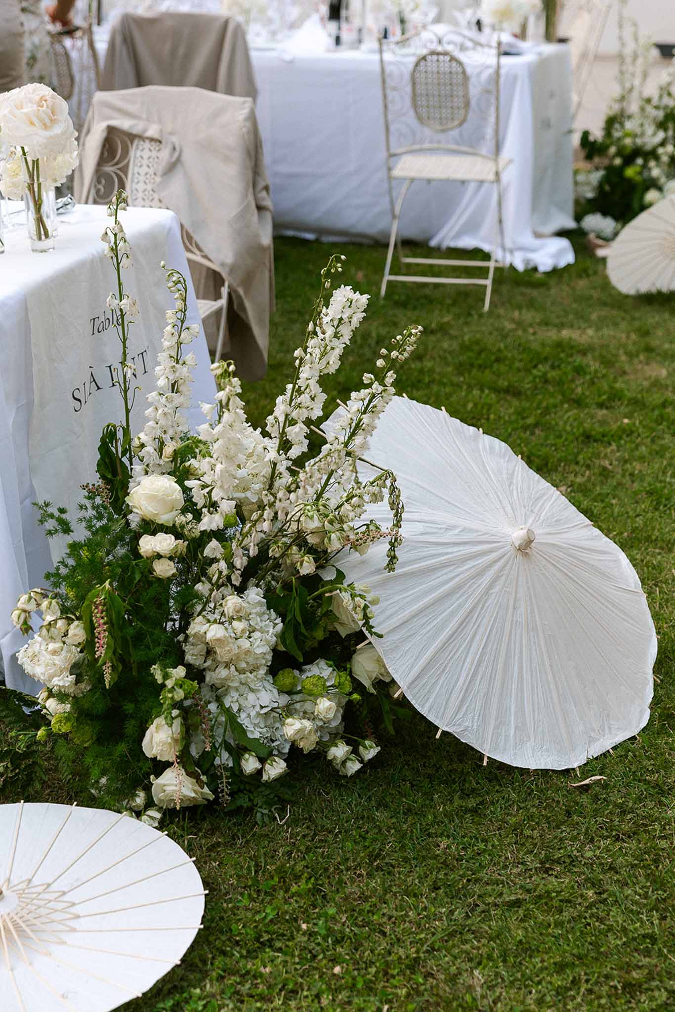 White delphinium and hydrangea ground arrangement with parasols beside table set with ivory rose bud vases on lawn