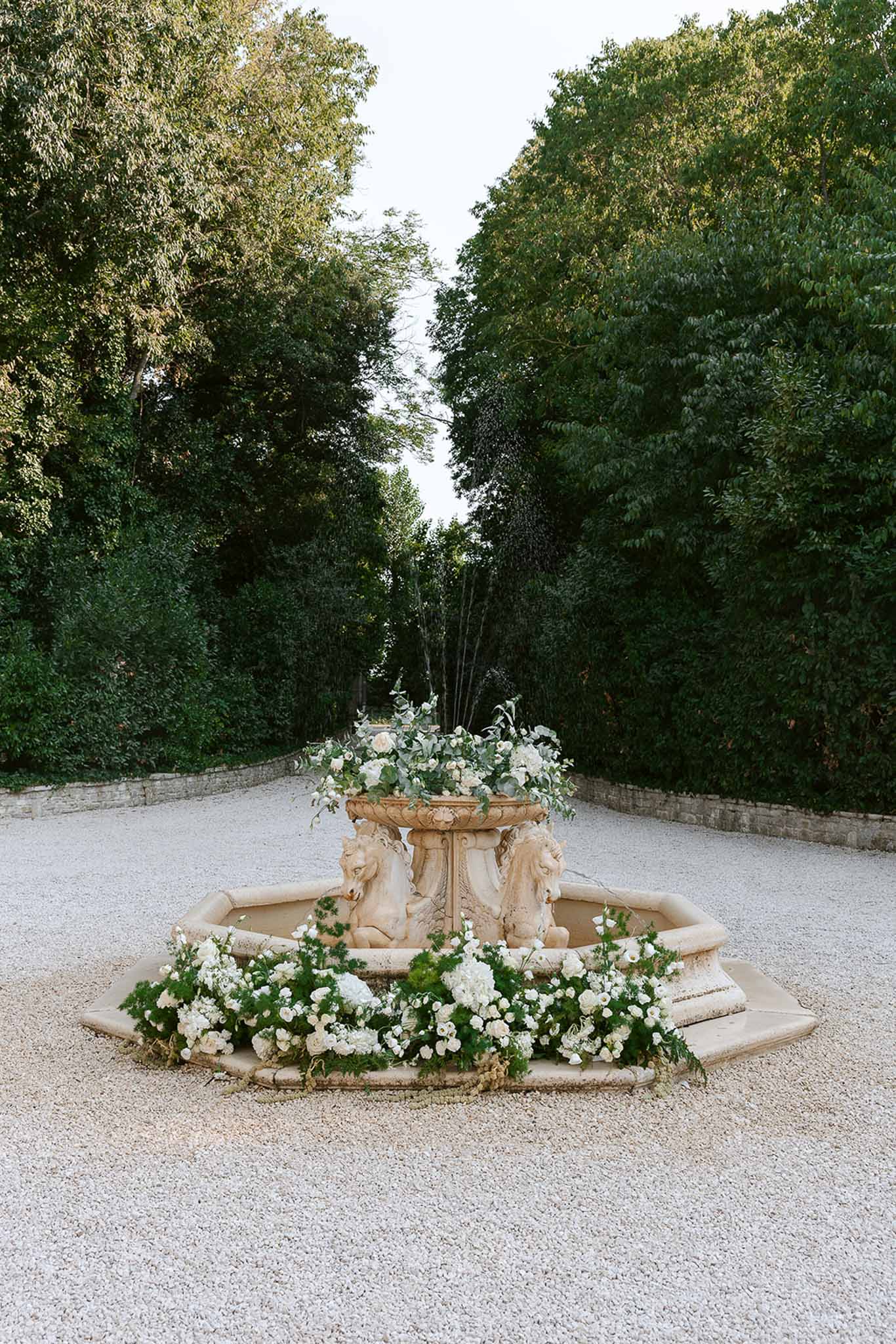 Stone fountain decorated with white roses and hydrangeas surrounded by green foliage in chateau gravel courtyard